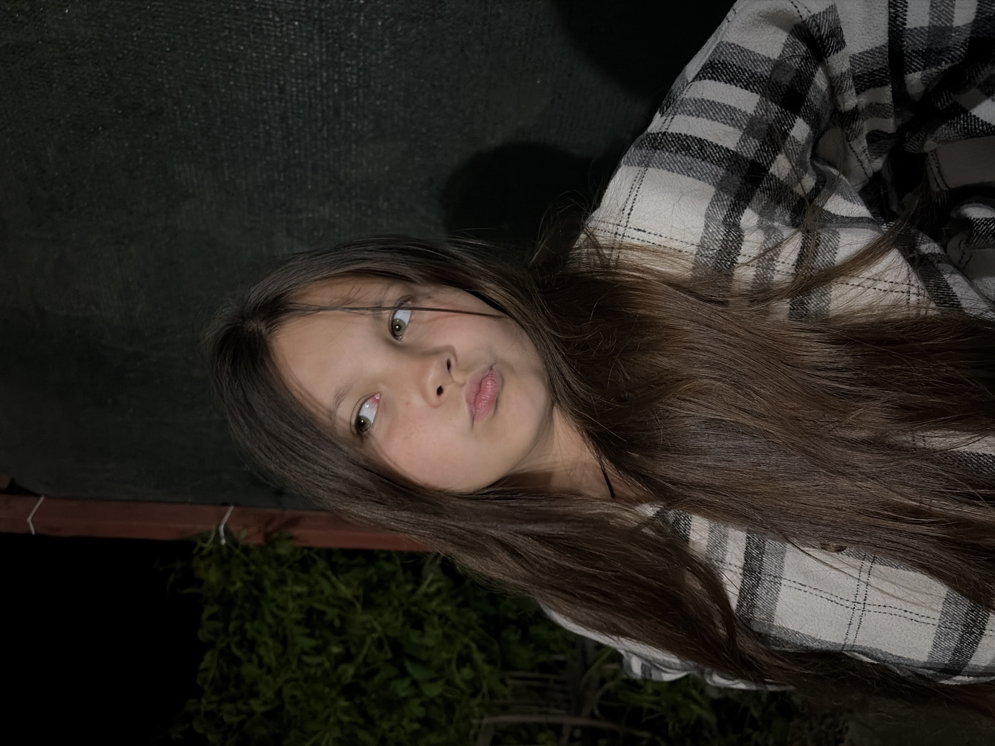 A young girl with long, brown hair and fair skin is posing for a selfie. She is wearing a black and white plaid shirt. The background is dark with some green foliage visible at the bottom left and a dark, possibly textured, surface behind her. The location is unspecified.