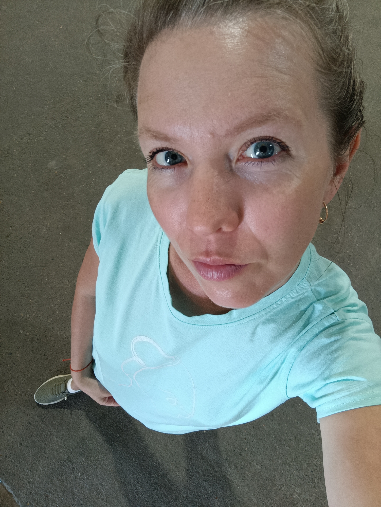 A high-angle full shot shows a woman with fair skin and blue eyes, looking upwards toward the camera. She has light-colored hair pulled back, revealing a gold hoop earring. She wears a light blue t-shirt with a white design. Her left hand rests near her hip. Her left foot is visible, clad in a grey athletic shoe with white laces. The background consists of grey pavement. There is no indication the image was taken in Chaikovsky, Russia.