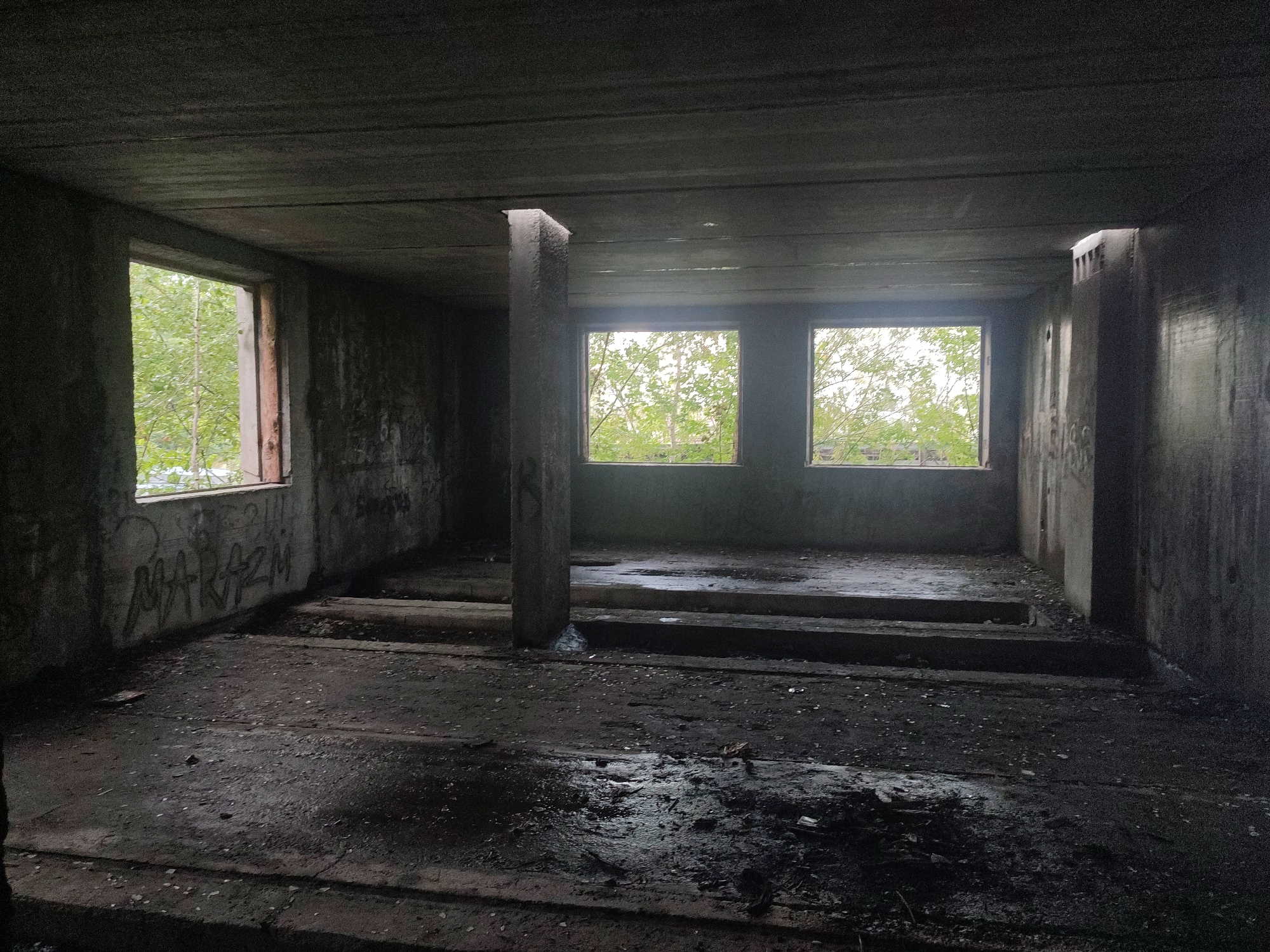 An interior view of an abandoned concrete building. Three windows are visible, two on the right wall and one on the left wall, all looking out onto lush green trees. The interior is dark and appears to be in a state of disrepair, with concrete walls and floors. Graffiti is visible on the walls, and puddles of water are present on the floor. A central concrete pillar supports the ceiling. This building is located in Novoaltaysk, Russia.