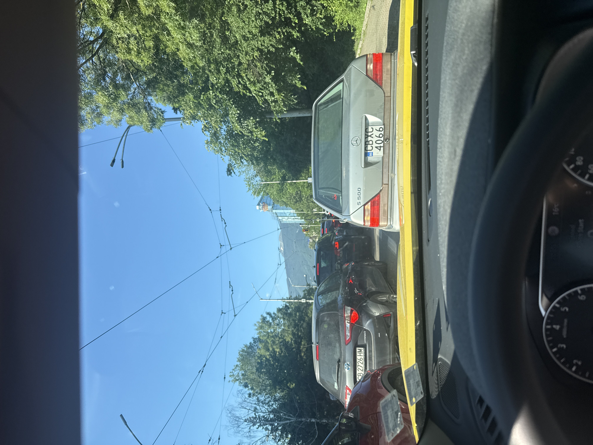 A street-level, eye-level, medium shot captures a traffic jam on a sunny day in Sofia, Bulgaria. The perspective is from inside a vehicle, looking forward through the windshield. A line of cars is stationary, indicating a traffic delay. In the foreground, the yellow hood and dashboard of the vehicle the photo is taken from are visible. Beyond that, several other cars are lined up, including a silver Mercedes S500 with license plate CBXC 4066, a dark gray Toyota Yaris hybrid with license plate CB 2726 HM, and a red vehicle. Overhead, electrical wires for what appear to be trams or trolleybuses crisscross the sky, along with streetlights. Lush green trees line the right side of the road, and in the distance, mountains can be seen under a clear blue sky. A modern glass building is also visible among the trees on the left side of the road. The scene suggests a typical urban commute in Sofia.