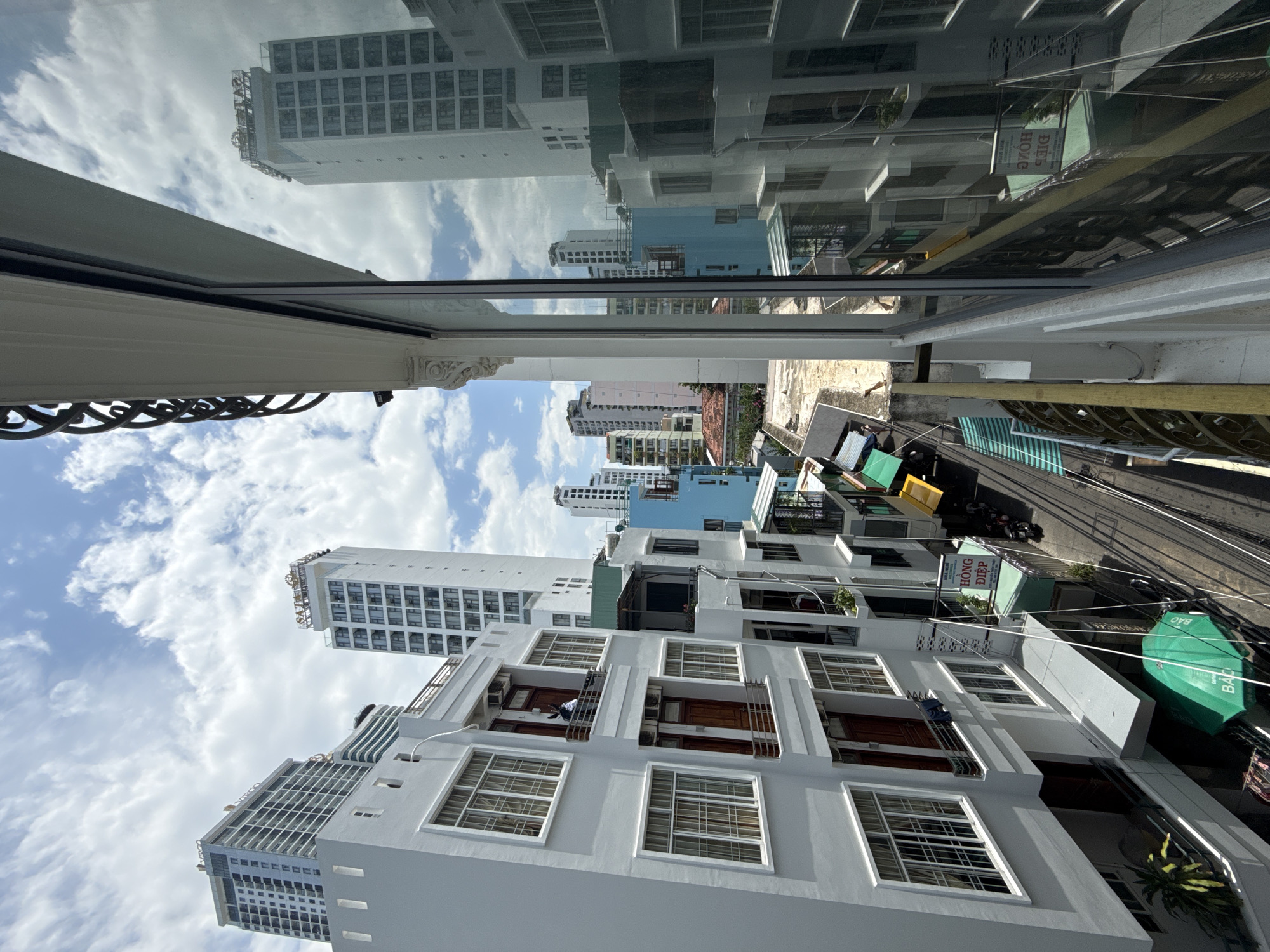 This is a high-angle, eye-level shot of a street scene in Nha Trang, Vietnam, dominated by modern architecture and a vibrant sky. The perspective is from a balcony or window of a building, looking down onto a street lined with multi-story buildings.
In the foreground, the edge of a white building with a decorative black metal railing is visible, framing the view to the right. A large window on this building reflects the sky and opposite buildings.
The main focus is the street below, which is a narrow thoroughfare with a paved surface. On either side of the street are buildings of varying heights and architectural styles. Many of them are white or light-colored, with numerous windows and balconies. Some buildings have a more contemporary design with large glass facades, while others appear older with more traditional window frames.
Several signs are visible on the buildings, some with Vietnamese text. For instance, a green sign with white lettering reads "HONG DIEP" which is likely a business name. There are also yellow awnings over some storefronts.
The street is somewhat busy, with a few motorcycles parked along the side. Power lines are strung overhead, crisscrossing the view. On the lower left, a large green umbrella is open, providing shade.
The sky is a prominent feature, filled with a mix of white, fluffy clouds against a bright blue backdrop. The sunlight creates shadows on the buildings and the street, indicating it is daytime. The reflections in the window in the foreground show more of the cityscape and the sky.
The image captures a typical urban environment in a coastal city like Nha Trang, characterized by a blend of residential and commercial buildings, busy streets, and a clear sky. The overall impression is one of a bustling and dynamic city.