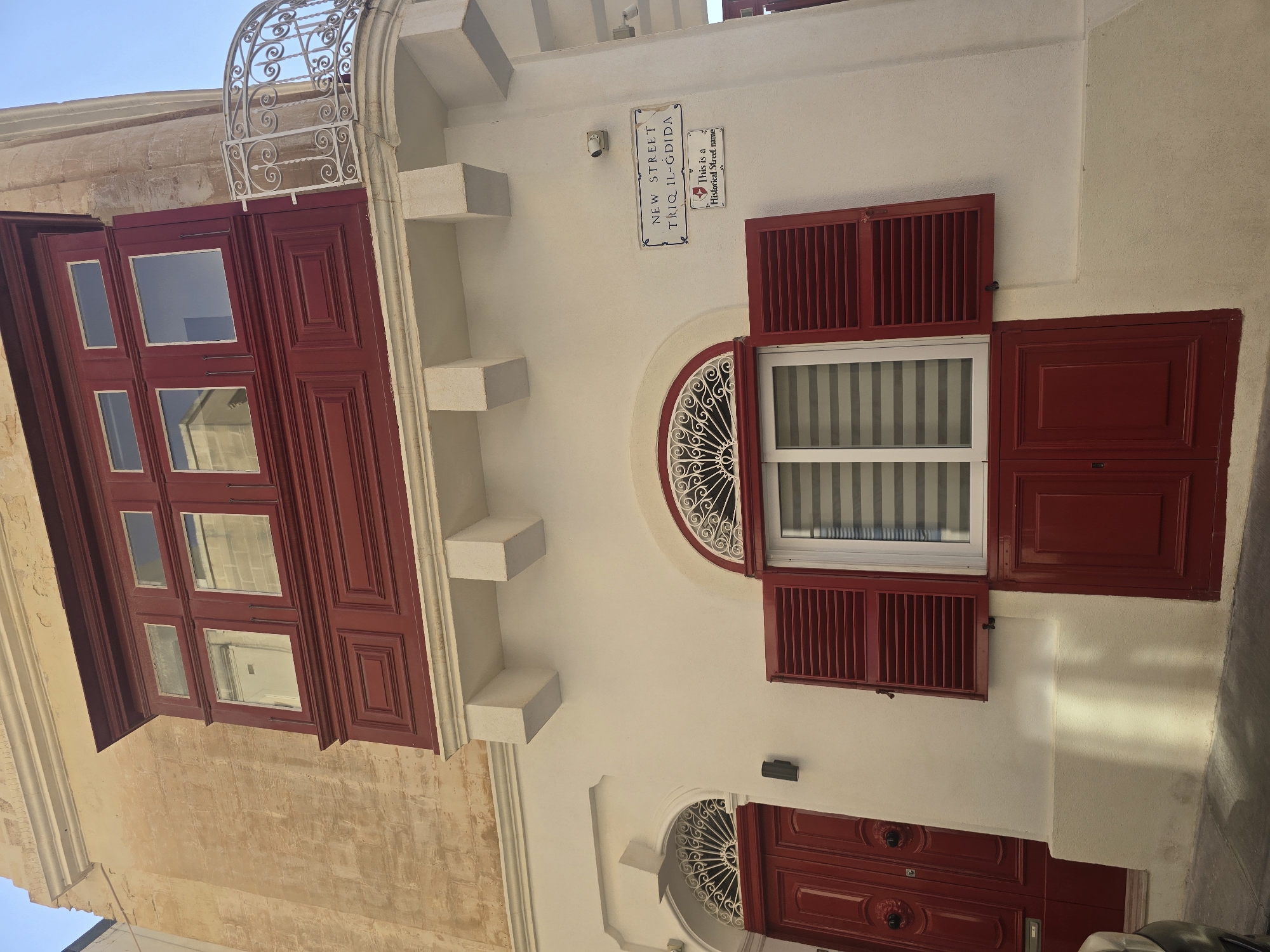 The image shows a facade of a building in Sliema, Malta, featuring a closed red wooden balcony with three windows at the top and paneled sections below. Adjacent to the balcony is a decorative white metal railing. The building is painted white, with a textured, light brown stone wall on the left side. There is a red wooden door with decorative carvings on the left, and a large, white-framed window with red shutters, partially open, in the center. Below the window is another red wooden door. A street sign indicates "NEW STREET TRIQ IL-GDIDA" and a smaller sign below it reads "This is a Historical Street name". A security camera is visible on the white wall above the street sign. The sky is clear and blue.