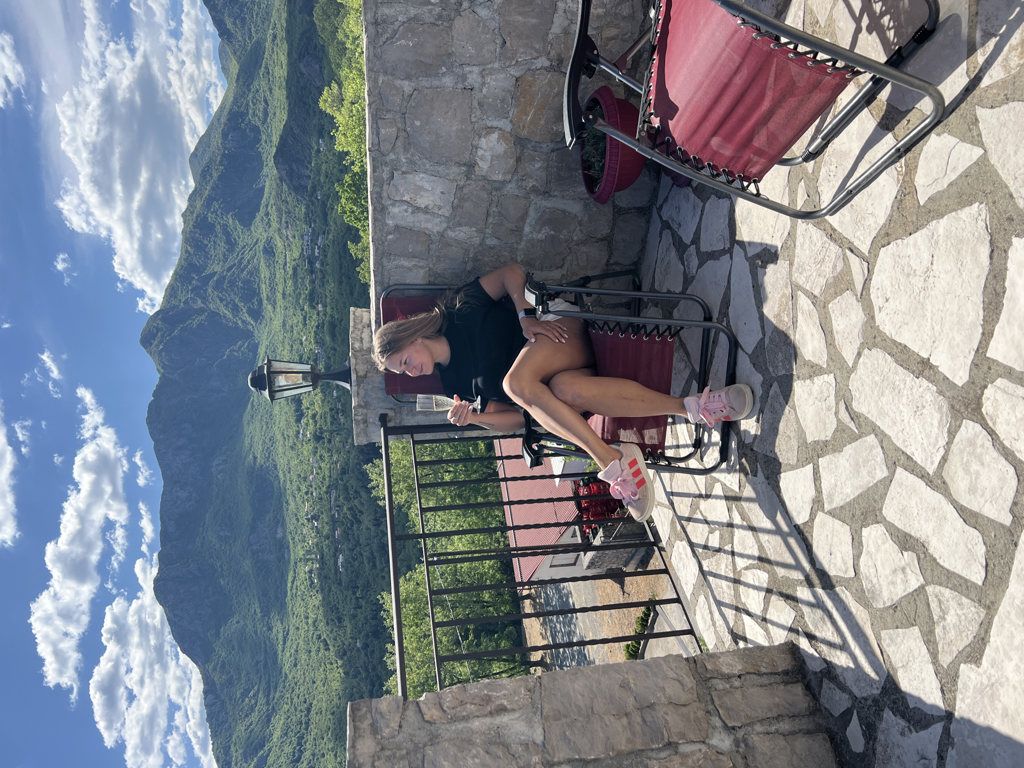 The image, originally rotated 90 degrees clockwise, captures a peaceful outdoor scene on a stone patio in Sotonići, Montenegro. A woman with medium-length brown hair is relaxing in a red fabric recliner chair, holding a champagne flute. She is dressed in a black t-shirt, light-colored shorts, and pink and white sneakers, with "adidas" visible on the side of her shoes. She appears to be gazing out at the view.
The patio is paved with irregular light-colored stone tiles. A low stone wall and a black metal railing form the boundary, with a lantern affixed to the wall. Beyond the railing lies a dramatic mountainous landscape; steep, verdant peaks covered in lush trees dominate the background. A few distant structures or a small settlement are nestled within the greenery. The sky above is a vibrant blue, adorned with scattered white cumulus clouds.
Strong sunlight creates distinct, elongated shadows from the chairs and railing across the patio, indicating it is either mid-morning or late afternoon. Another red recliner chair and a red potted plant are partially visible. The scene conveys a sense of tranquility and enjoyment of the stunning natural surroundings.