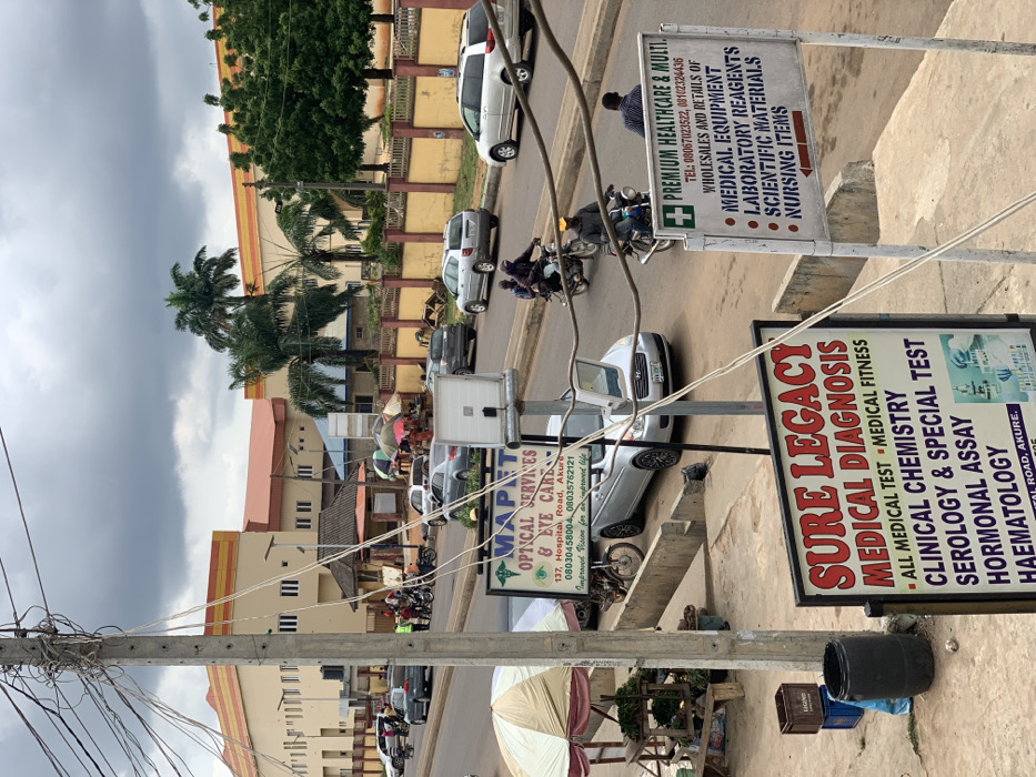 The image shows a street scene in Akure, Nigeria, at the coordinates 7.241 latitude and 5.195 longitude. The street is lined with buildings and palm trees. There are several vehicles parked along the street and some in motion. There are people walking on the sidewalk. In the foreground, there are two signs, one for "MAPET OPTICAL SERVICES & EYE CARE" and one for "SURE LEGACY MEDICAL DIAGNOSIS." The sign for "SURE LEGACY MEDICAL DIAGNOSIS" advertises "ALL MEDICAL TEST" including "CLINICAL CHEMISTRY," "SEROLOGY & SPECIAL TEST," "HORMONAL ASSAY," and "HAEMATOLOGY." The sign for "MAPET OPTICAL SERVICES & EYE CARE" includes a phone number and the address "137, Hospital Road, Akure". The street is paved and has a curb along the side of the road. There is a trash can on the sidewalk. The sky is overcast.
