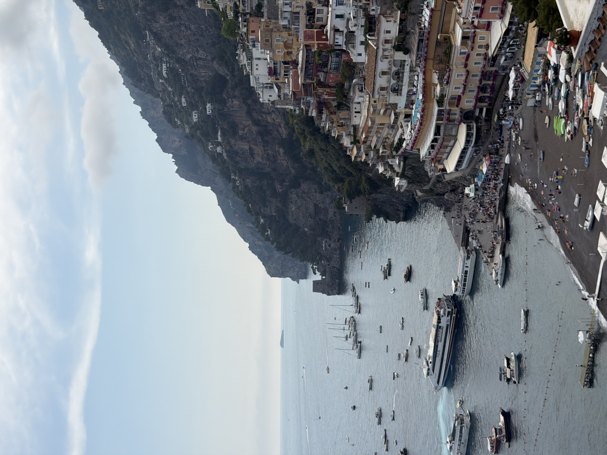 This image depicts the coastal town of Positano, Italy. The town's colorful buildings cascade down a steep cliff towards the sea. A dark, rocky mountain forms a backdrop. The sea is filled with boats, ranging from small sailboats to large ferries. A beach is visible at the foot of the town, crowded with people and boats. The sky is partly cloudy with bright light from the sun.
