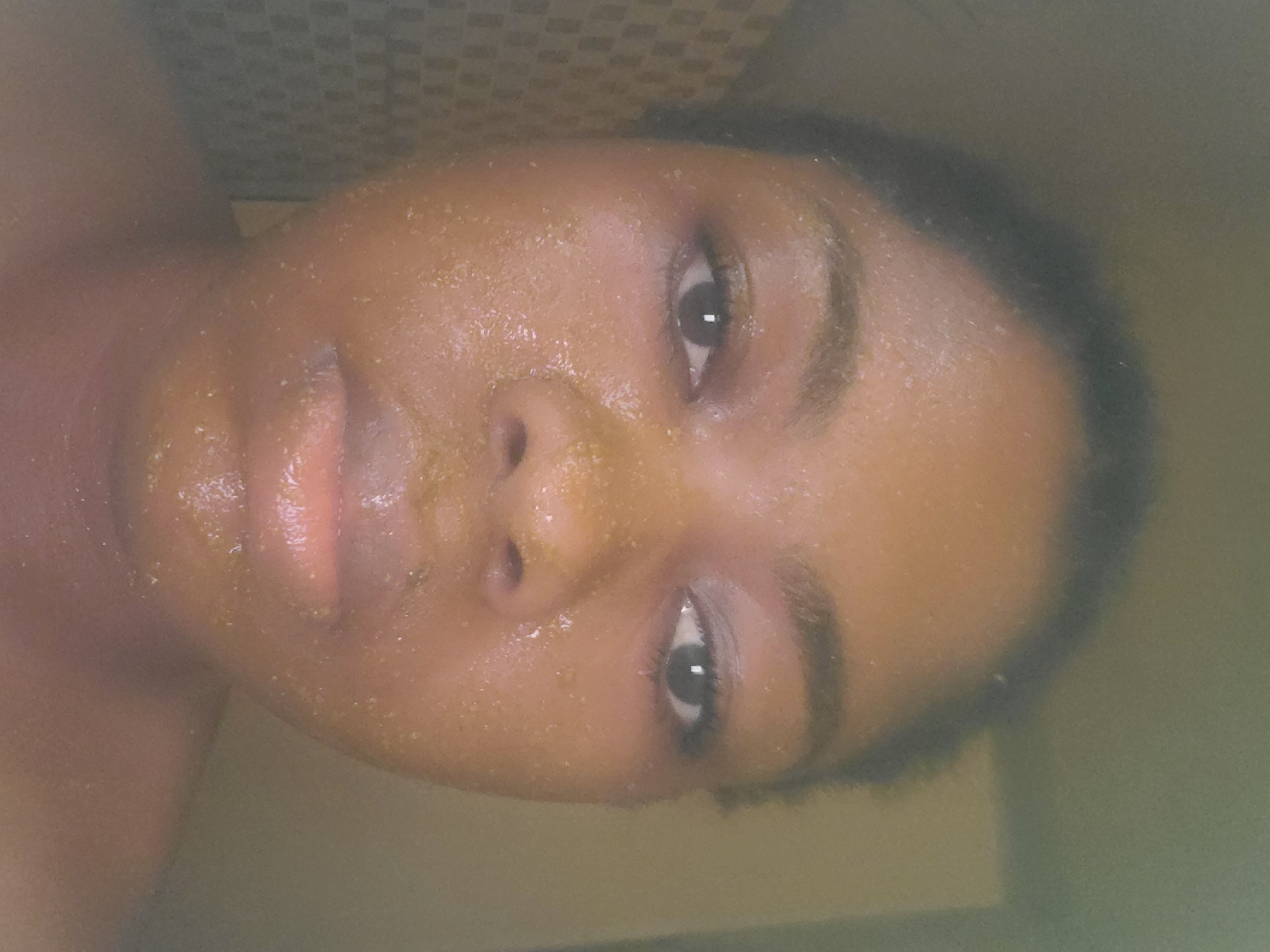 The scene is set in Kakau, Nigeria. A young adult individual with dark skin is depicted in a close-up portrait, facing directly towards the viewer. The person's face is thoroughly covered with a granular, yellowish-brown substance, consistent with a facial mask or scrub, exhibiting small, distinct particles across the forehead, cheeks, nose, and chin. Dark hair is pulled back from the face. The individual has dark, defined eyebrows, and dark eyes that engage directly with the camera. The lips are full and display a natural, moisturized appearance. The shoulders and neck are visible, suggesting the person is bare-chested or wearing a strapless top. The background on the left features a tiled wall with a dark, square grid pattern. The right side of the background is a lighter, uniformly colored wall or surface. The lighting is soft and appears to originate from the front, illuminating the face.