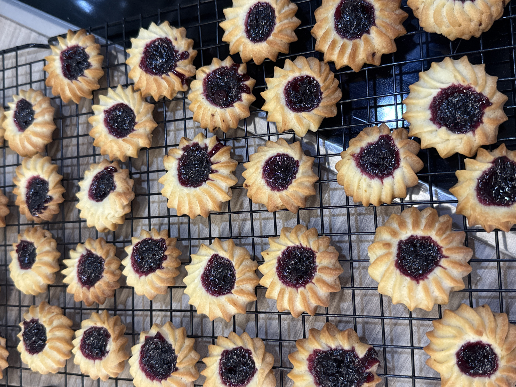 The image shows a cooling rack filled with approximately 28 jam-filled cookies. The cookies are circular with a fluted edge, creating a starburst pattern, and have a dark, jam-like filling in the center. The cookies are arranged in neat rows on the black wire cooling rack. The background is out of focus. There is no indication of Krasnoyarsk, Russia.