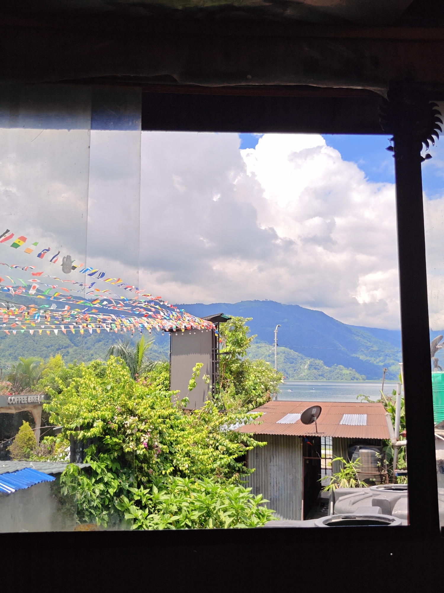 This image captures a scenic view from an elevated vantage point in Pokhara, Nepal, likely overlooking Phewa Lake. In the foreground, a series of colorful international flags are strung across a wire, creating a festive canopy over what appears to be a cafe or restaurant area. The flags are small and triangular, displaying various national symbols. Below the flags, lush green foliage dominates the middle ground, with trees and bushes laden with leaves. Peeking through the greenery are glimpses of structures, including a corrugated metal roof with a satellite dish. A sign that says "COFFEE BREAK" is visible on the left. In the distance, a serene lake stretches out, bordered by rolling green hills that ascend into majestic mountains, their peaks softened by a layer of atmospheric haze. The sky above is a dynamic mix of bright blue patches and voluminous white clouds, suggesting a pleasant, possibly partly cloudy day. The overall impression is one of a relaxed, tourist-friendly environment with a breathtaking natural backdrop.