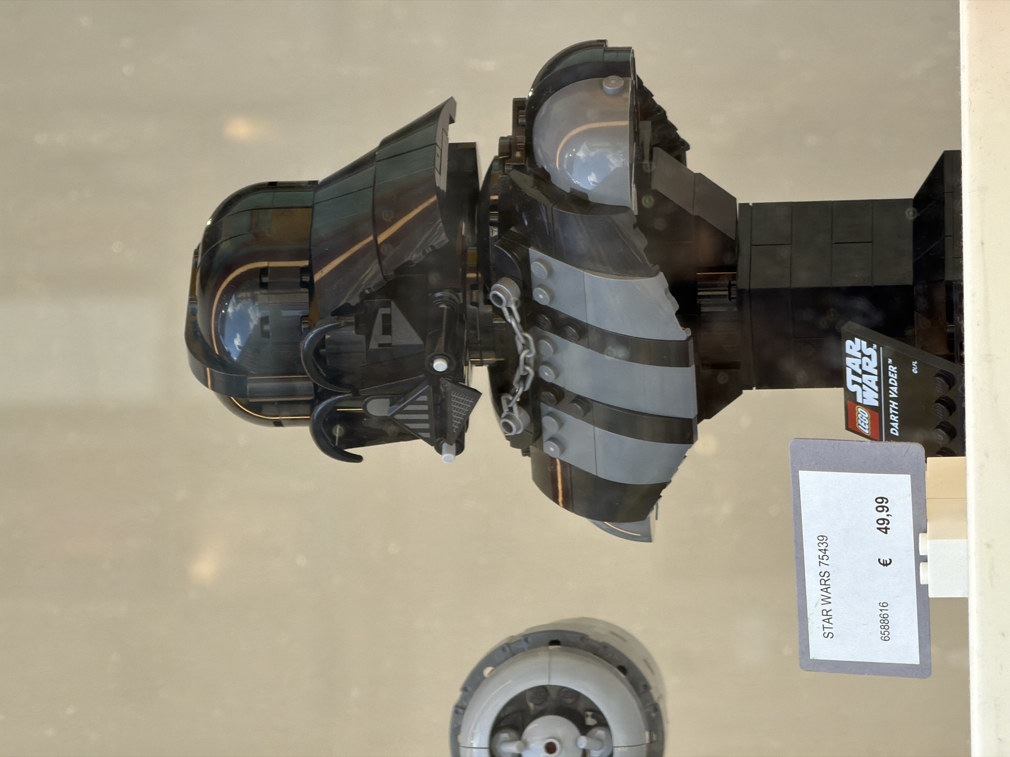An image captured in Milan, Italy, displays a LEGO Star Wars Darth Vader buildable bust, presented vertically within what appears to be a retail or exhibition display case. The main subject is the intricately constructed model, predominantly in black and dark grey LEGO bricks, featuring Darth Vader's helmet and upper chest armor. Some parts of the model have a shiny finish, reflecting ambient light and hints of the surrounding environment. A small, black information plaque attached to the base of the model is visible, bearing the "LEGO" logo, "STAR WARS™", and "DARTH VADER™" branding, along with "©LFL".
Attached to the right side of the display is a grey price tag, positioned horizontally. The text on the price tag reads "STAR WARS 75439" at the top, "€ 49,99" in the middle, indicating the price in Euros, and "6588616" at the bottom, likely a product or inventory number. The background is a plain, light, neutral color, suggesting a minimalist display setting. The overall presentation indicates a product available for sale, possibly in a store window or an in-store display, showcasing the LEGO set's design and detail. The image itself is rotated 90 degrees counter-clockwise.
