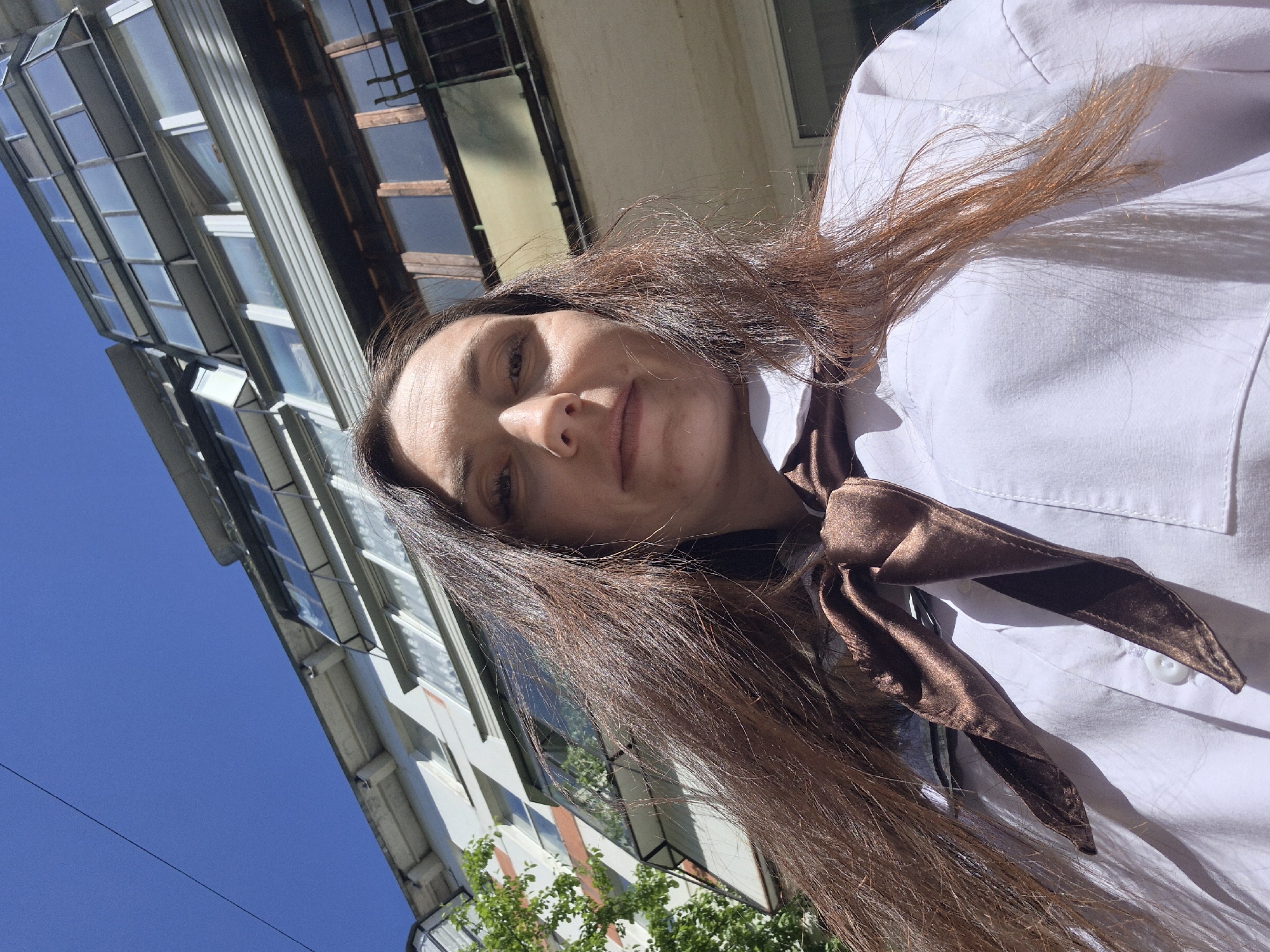 A woman with long brown hair and a white shirt with a brown necktie stands outside in a city. The background shows a building with a modern facade and large windows. The sky is clear and blue. The location appears to be a city street, possibly in Unknown, Unknown, judging by the architectural style.
