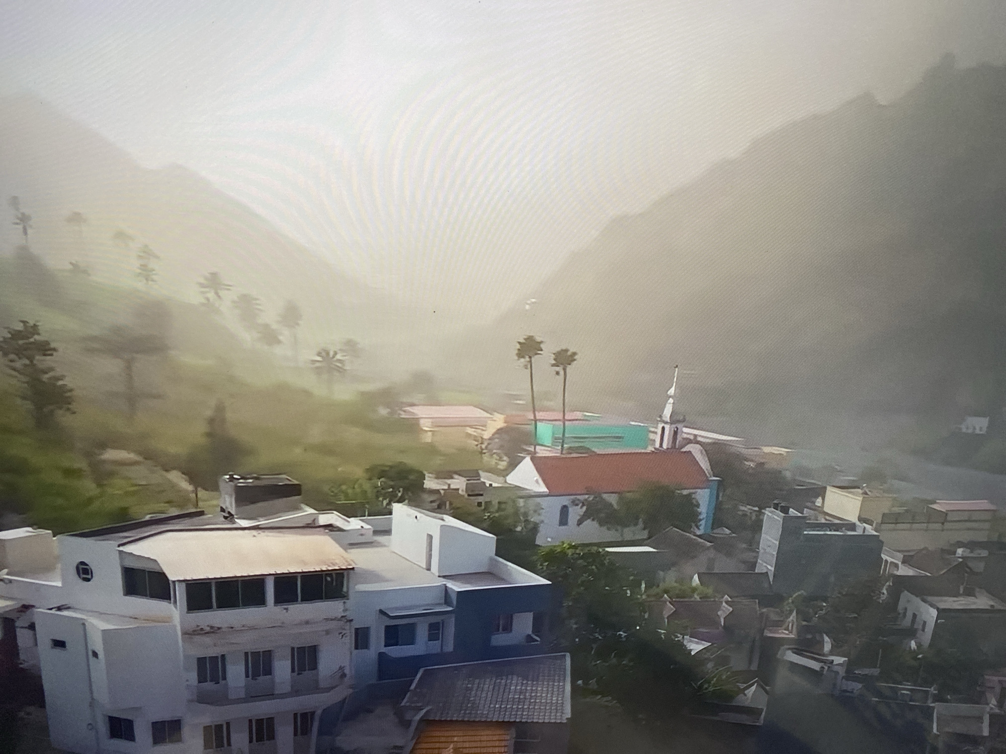 This is an aerial, daylight, wide-angle shot of a small village nestled in a valley. The village consists of several buildings with pitched and flat roofs in various colors, including white, red, teal, and yellow. Some buildings appear to be residential, while one building has a steeple, suggesting it might be a church. Palm trees are visible scattered throughout the scene. The village is surrounded by green, mountainous terrain that is partially obscured by mist. The sky is overcast with bright, hazy conditions. The scene is static and there are no people or animals visible. The location is not in Minsk, Belarus.