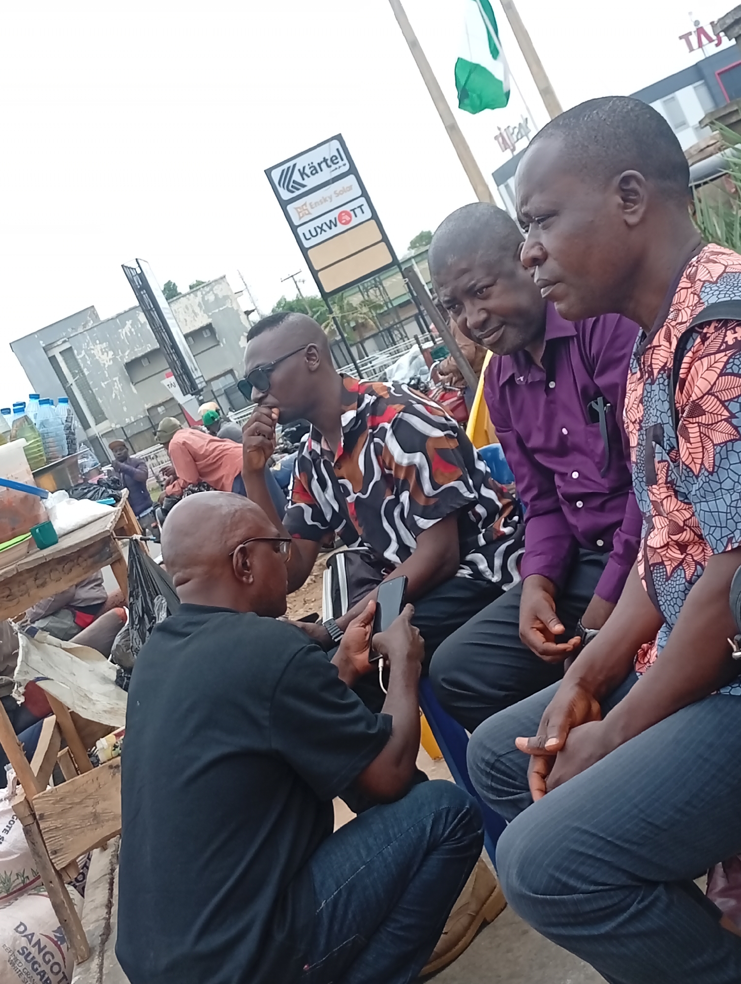 The image shows four men in an urban environment, possibly the city of Maiburiji, Nigeria. They are positioned near a busy street, with a partially obscured Nigerian flag and various commercial signs visible in the background, including those for "Kärtel", "Ensky Solar", "LUXWOTT", and "Taj". A man in a black shirt is holding a mobile phone in front of him, seemingly showing something to the others. To his left, a man with sunglasses is sitting and possibly eating something. Two other men are sitting beside him, one in a purple shirt, the other in a patterned shirt. They are all looking towards the phone. The setting appears to be a roadside gathering, with additional people and goods like water bottles and sugar visible in the background, suggesting a bustling marketplace or waiting area. Some of the men are sitting on blue chairs.