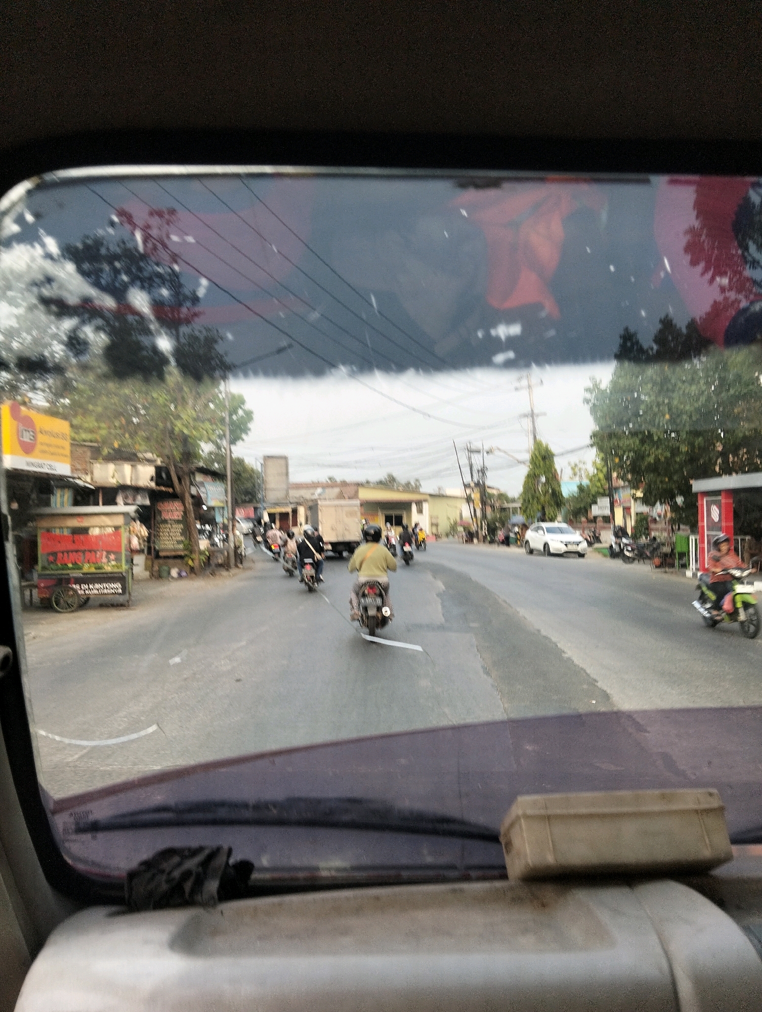 The image depicts a street scene in Semarang, Indonesia, viewed from the interior of a vehicle. The foreground shows the dashboard and windshield of the vehicle, with a dusty object placed on the dashboard. The windshield is dirty and reflects the sky, trees, and some red objects. The middle ground shows a street with several motorcycles and a white car. There are also roadside structures, including small shops and stalls, and trees lining the street. The background shows more buildings and utility poles. The lighting suggests it is daytime, with an overcast sky.