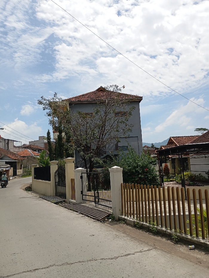 The scene is a residential street in Kecamatan Kutawaringin, Indonesia (latitude: -6.995, longitude: 107.539). There is a two-story house with a gray facade and a red tile roof. The house is surrounded by a black metal fence and a low wall. There is a small gate on the left side of the house. The house is set back from the street, which is paved in gray concrete. On the right side of the street, there is a fence made of vertical wooden slats. Behind the fence, there is a second house with a brown tile roof and a covered patio. There is a green lawn in front of the second house. The sky is blue with white clouds. There is a lone motorcycle parked on the left side of the street, indicating that there may be a resident nearby. The scene is quiet and peaceful, with no people or animals in sight.