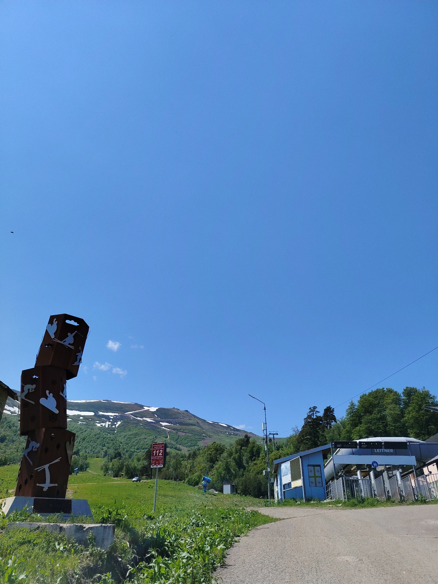 This is an outdoor, daytime, wide-angle photograph taken on a clear, sunny day. The scene depicts a mountain resort area, likely in Bakuriani, Georgia, given the presence of a ski lift. In the foreground to the left is a large, abstract sculpture made of stacked, dark brown metal cubes. Each cube features cut-out silhouettes of various winter sports figures, such as skiers and snowboarders, as well as hearts and snowflakes. The sculpture is mounted on a concrete base. To the right of the sculpture is a paved road leading into the scene. Further back on the right is a blue building with a Leitner ski lift structure attached. The ski lift appears to be operational, with a covered cabin visible. The background is dominated by a lush green mountainside, dotted with trees. The upper slopes of the mountain are partially covered in snow, indicating a high altitude and possibly the tail end of a ski season or preparation for winter sports. A red and white sign with the number "112" is visible near the ski lift. A car is parked on the grassy area in the mid-ground. The sky is a vibrant, cloudless blue.