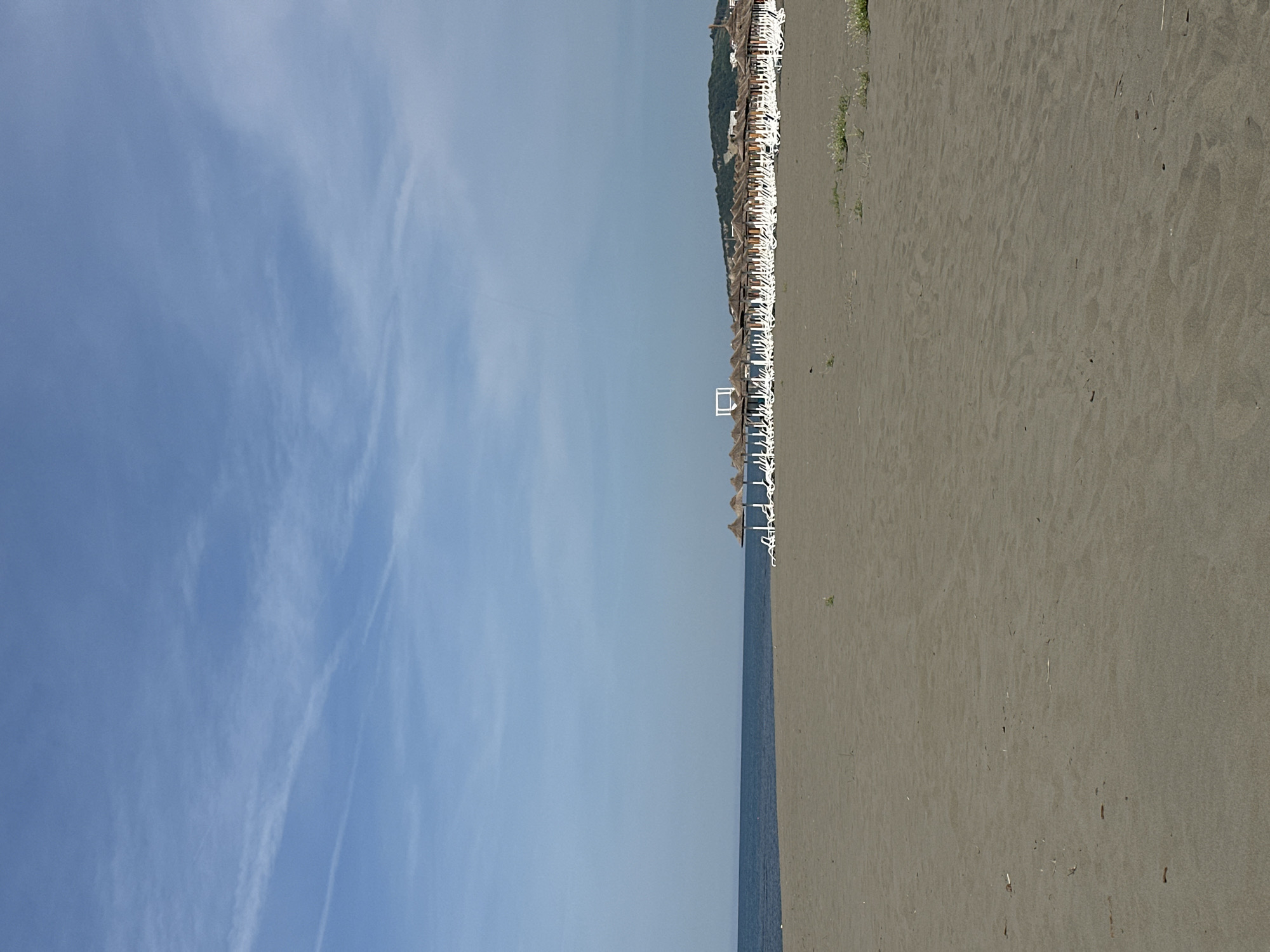 The image depicts a beach scene in Donji Štoj, Montenegro. The scene includes a wide sandy beach on the right, the Adriatic Sea in the center, and a predominantly clear sky with some clouds on the left. A row of beach umbrellas and chairs lines the edge of the beach, near the water. The sand is a light gray or tan color. The water appears calm.
