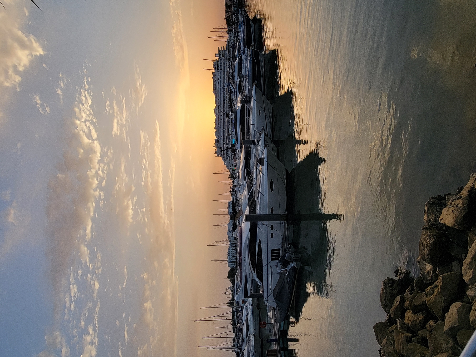 The image captures a serene sunset scene at the Quarteira Marina in Quarteira, Portugal. The sky is painted with warm hues of orange and yellow as the sun dips below the horizon, casting a golden glow over the water and the docked yachts. Numerous white yachts of various sizes are neatly lined up in the marina, their reflections shimmering on the calm water. In the background, a large, modern building complex stands against the colorful sky. The foreground shows a rocky embankment on the left side of the image, leading into the water. The overall atmosphere is peaceful and picturesque, highlighting the beauty of the coastal city at dusk.