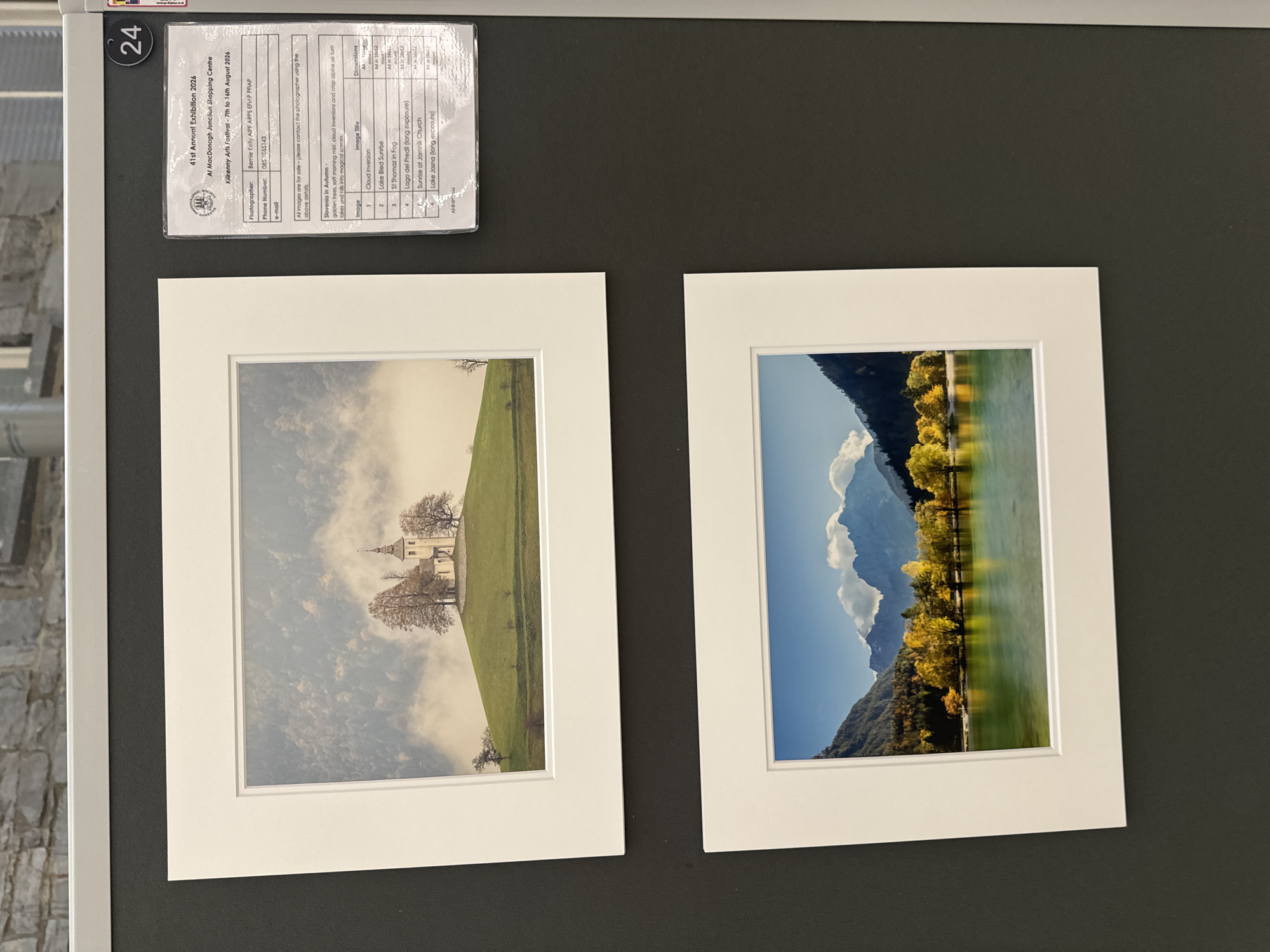 The image displays an exhibition of photographs at the MacDonagh Junction Shopping Centre in Kilkenny, Ireland. The exhibition is the 41st Annual Exhibition 2026, hosted by the Kilkenny Arts Festival, running from the 7th to the 16th of August. Two framed photographs are visible, mounted on a dark grey wall. The top photograph features a church on a grassy hill, partially obscured by mist and surrounded by trees. The bottom photograph depicts a lake with mountains and autumn foliage in the background. A printed information card is attached to the wall next to the top photograph, detailing the exhibition, photographer Bernie Kelly, contact information, and a list of the displayed images with their titles and dimensions. The number '24' is visible in the top right corner of the frame, likely indicating the exhibit number.