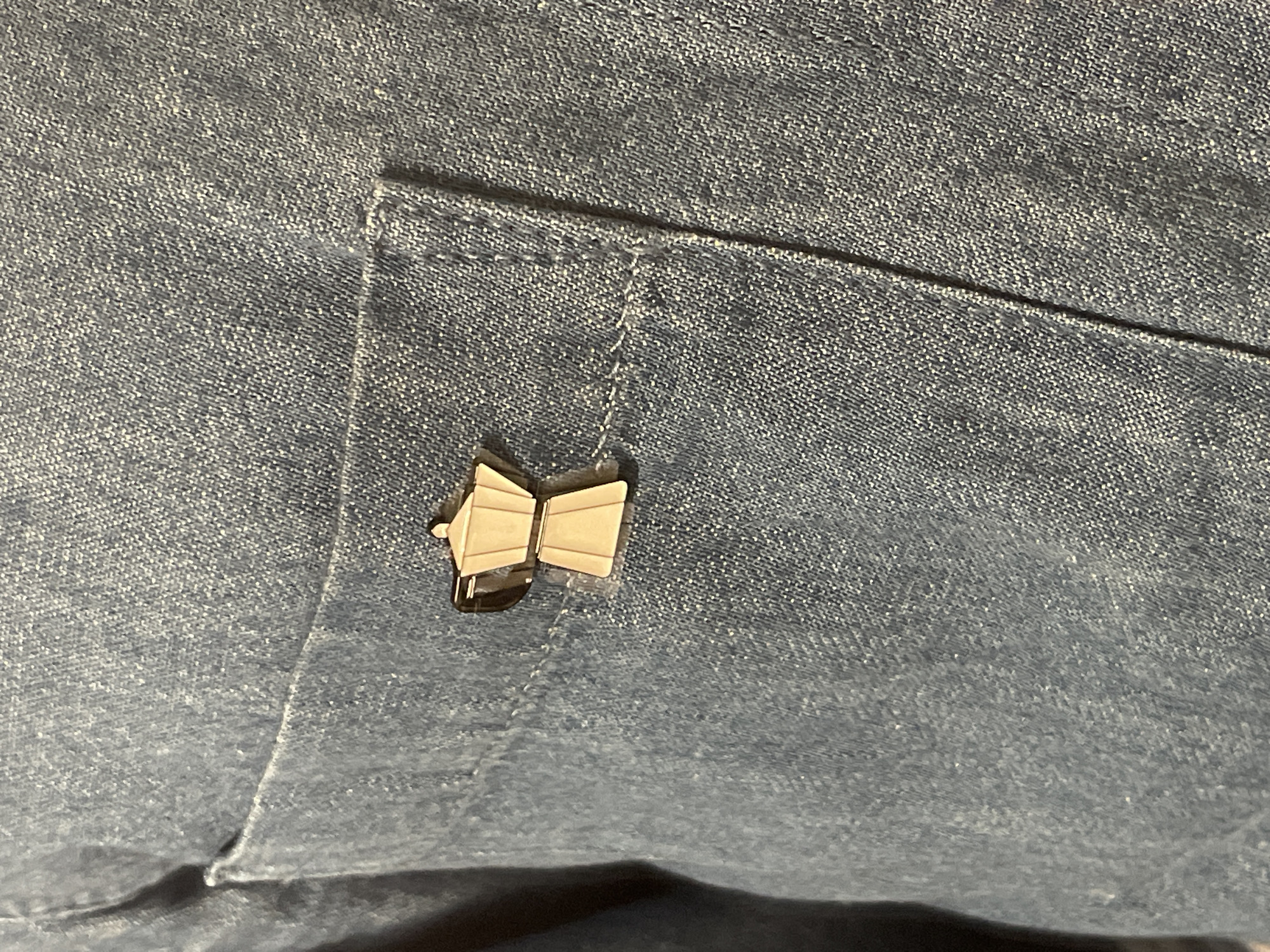 A close-up, eye-level shot focuses on a blue denim shirt pocket. A small, gold-colored lapel pin shaped like a moka pot is affixed to the pocket. The pin has two sections representing the coffee maker's base and top, with a dark outline emphasizing its form. The denim fabric shows a visible weave pattern and some light wear. The background is a continuation of the denim shirt. The location is not specified.