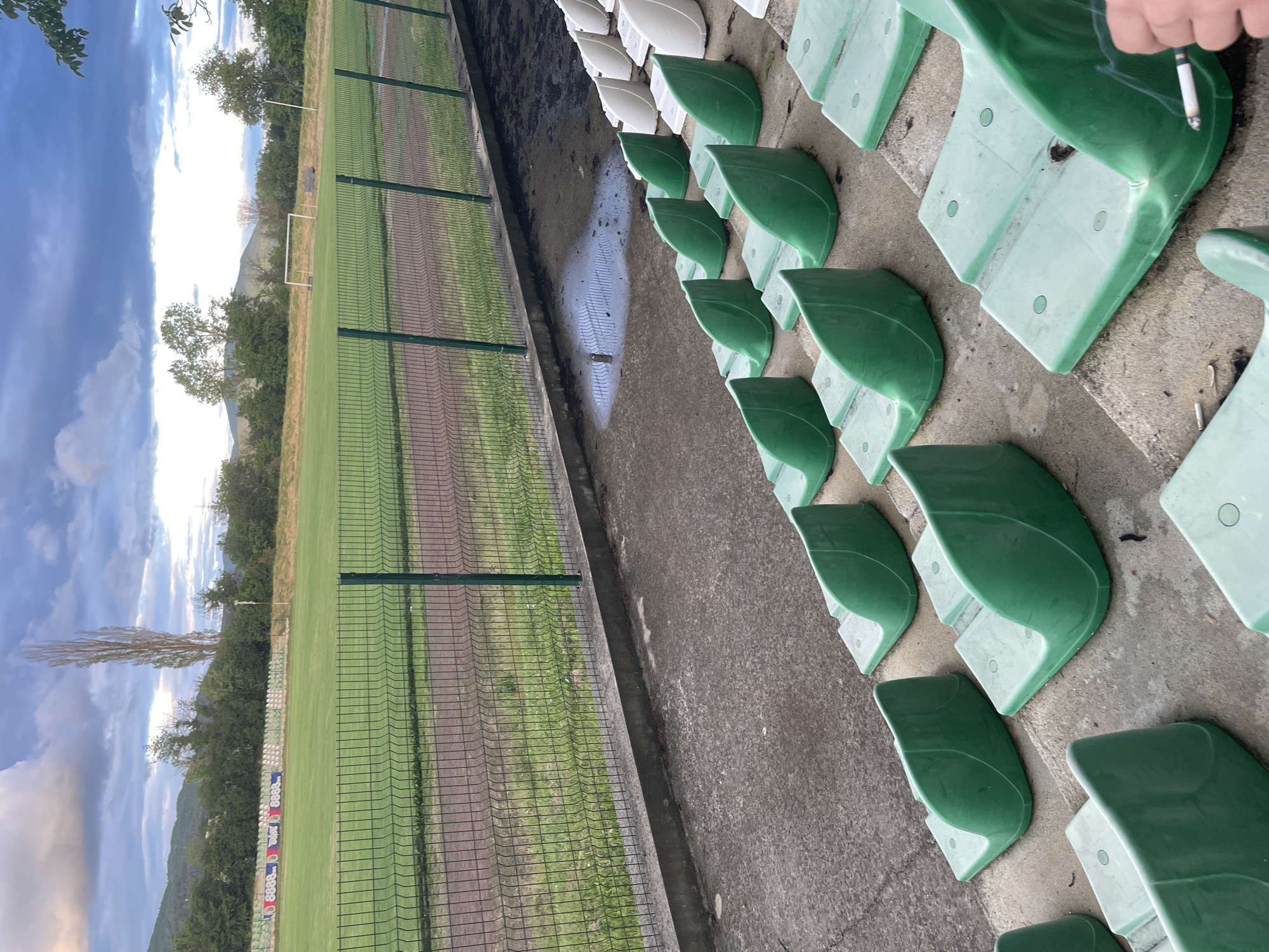 The image captures a view from the stadium stands overlooking a sports field, likely a football pitch, under a cloudy sky. The photograph is oriented vertically, showing the sky on the left and the stands on the right.
In the immediate foreground, rows of green and some white plastic stadium seats are visible on concrete steps. The concrete walkways are worn and marked with dirt and puddles of water, suggesting recent rain. Several discarded cigarette butts are scattered on the ground. A hand holding a lit cigarette with smoke wafting upwards is visible in the upper right corner of the frame.
Adjacent to the seating area, a drainage channel runs parallel to a tall chain-link fence that separates the stands from the playing field. Beyond this fence, the sports field stretches out, featuring areas of green grass and what appears to be a track or dirt patch. A white football goal stands prominently in the middle distance on the grass.
In the background, a line of trees fringes the field, with some appearing bare. Distant mountains or hills are visible beneath a dramatic, partly cloudy sky, featuring both bright white and darker grey clouds, suggesting either late afternoon or early evening light. On the far side of the field, behind another fence line, a banner or sign is partially visible, displaying numbers, notably "8888," along with other indistinct text or logos.
The scene depicts a quiet moment at a stadium in Ihtiman, Bulgaria, possibly between events, with only one person implied to be present in the stands.