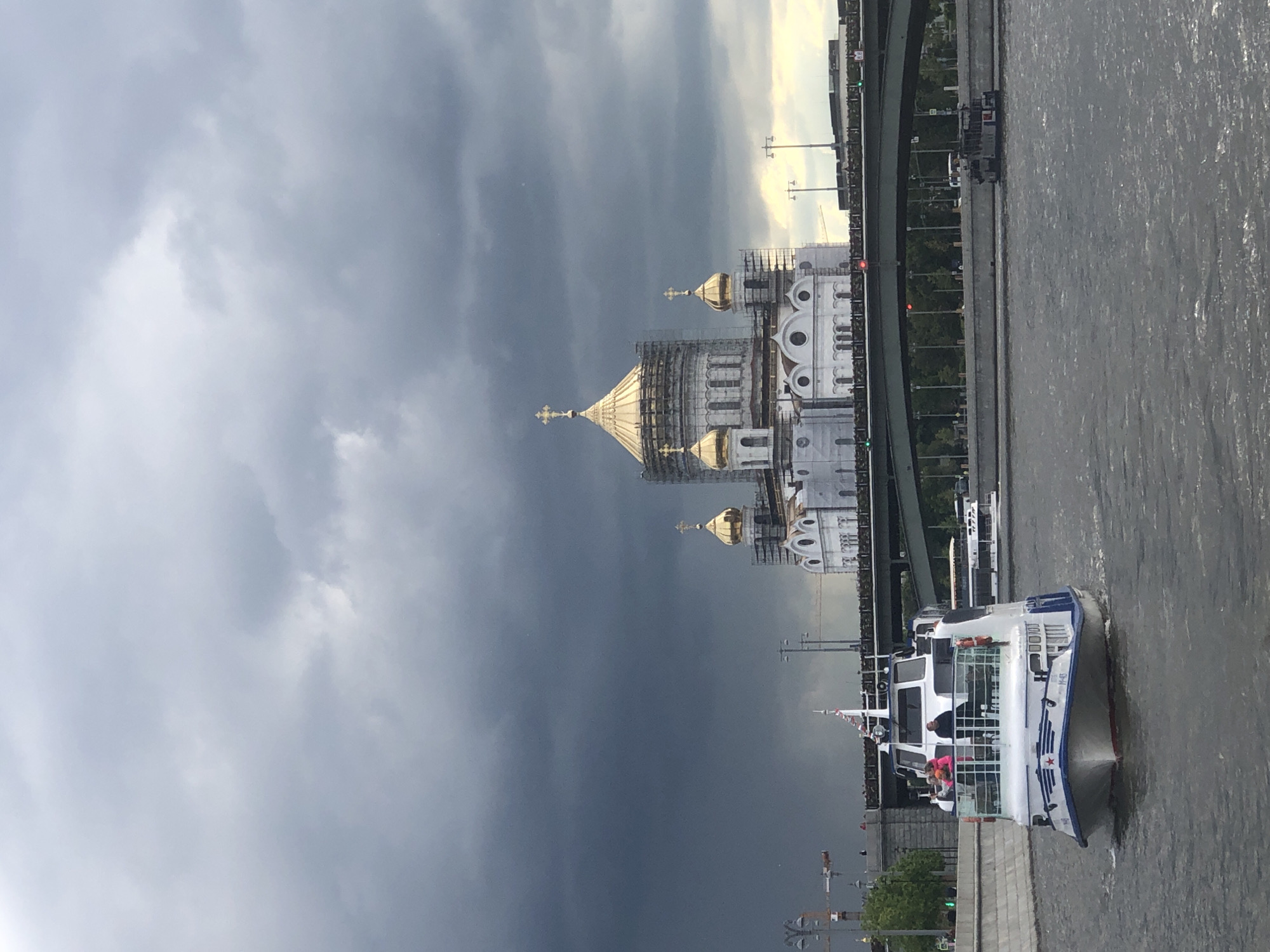 The image shows the Cathedral of Christ the Savior in Moscow, Russia, as seen from the river. The sky is overcast with dark storm clouds. The cathedral is white with gold domes, some of which are covered in scaffolding. A white boat is on the water near the riverbank. A bridge is visible to the right of the cathedral.