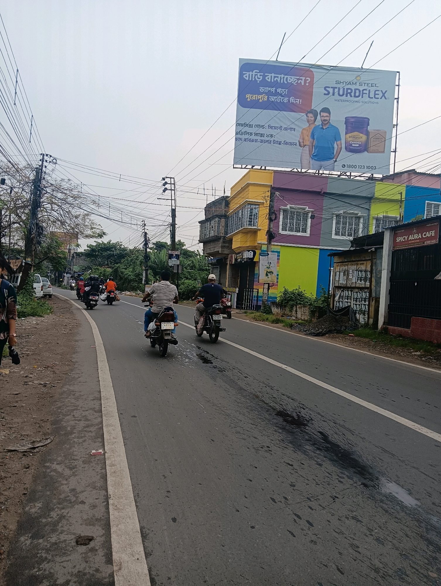 The image depicts a street scene in Barrackpore, India, on an overcast day. The road is paved and has a white line marking its center. Several motorbikes are visible on the road, with riders and passengers. In the foreground, a person is walking on the left side of the road, with their back to the camera. On the right side of the road, there are buildings. One building features a vibrant, multi-colored facade with yellow, purple, green, and blue sections. A billboard is mounted on top of this building, advertising "SHYAM STEEL STURDFLEX WATERPROOFING SOLUTIONS." Below the billboard, a sign reads "SPICY AURA CAFE." The environment appears to be urban, with trees and power lines visible. The road has some wet patches and dark stains.