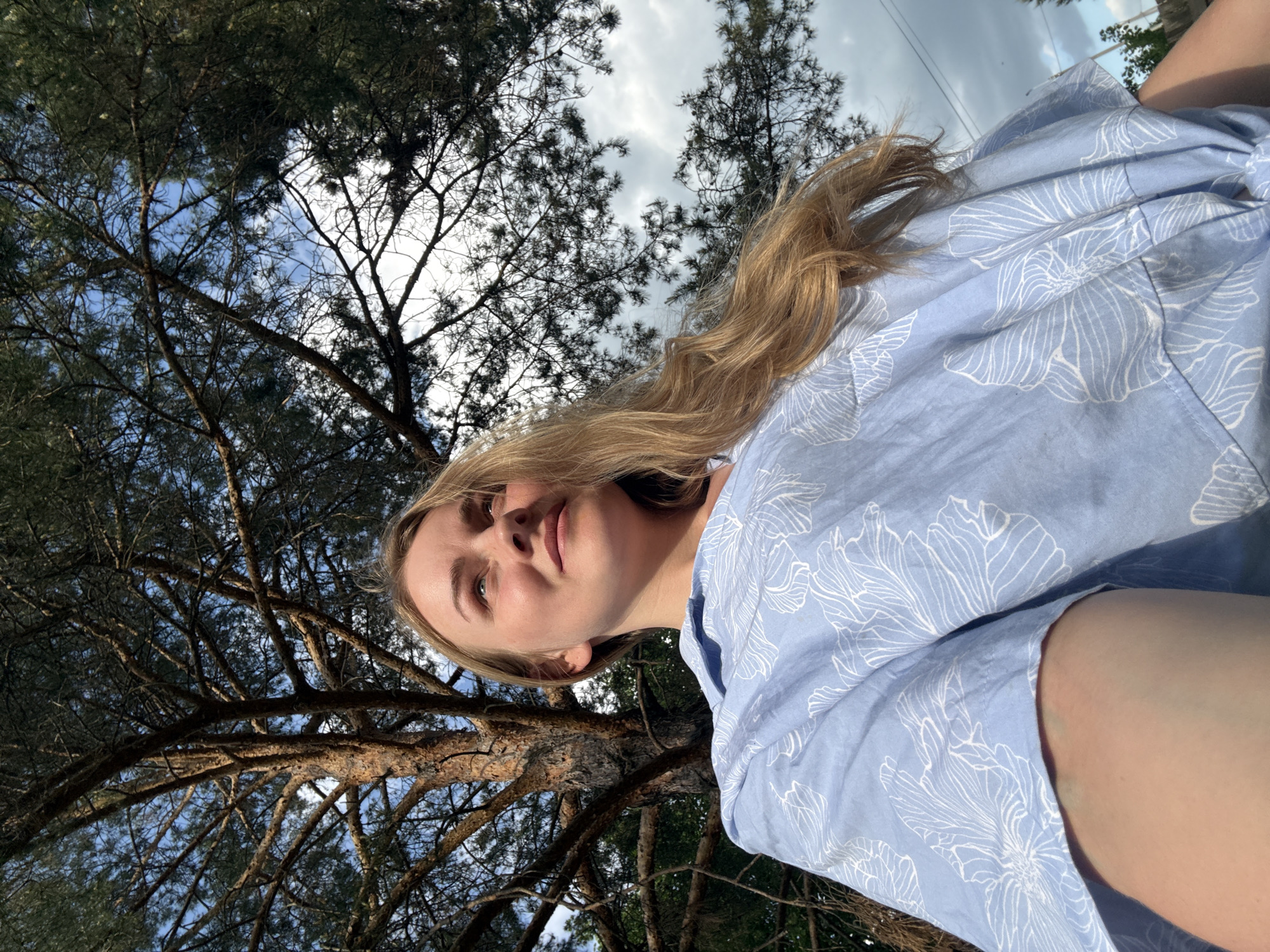 A young woman with blonde hair is captured from a low-angle perspective in what appears to be an outdoor setting, likely in a park or wooded area. She is wearing a light blue blouse with a white floral pattern. The background features a dense canopy of trees against a partly cloudy sky. The trees have slender, upward-reaching branches, suggesting they could be pine trees. The location is in the city of Chaikovsky, Russia.