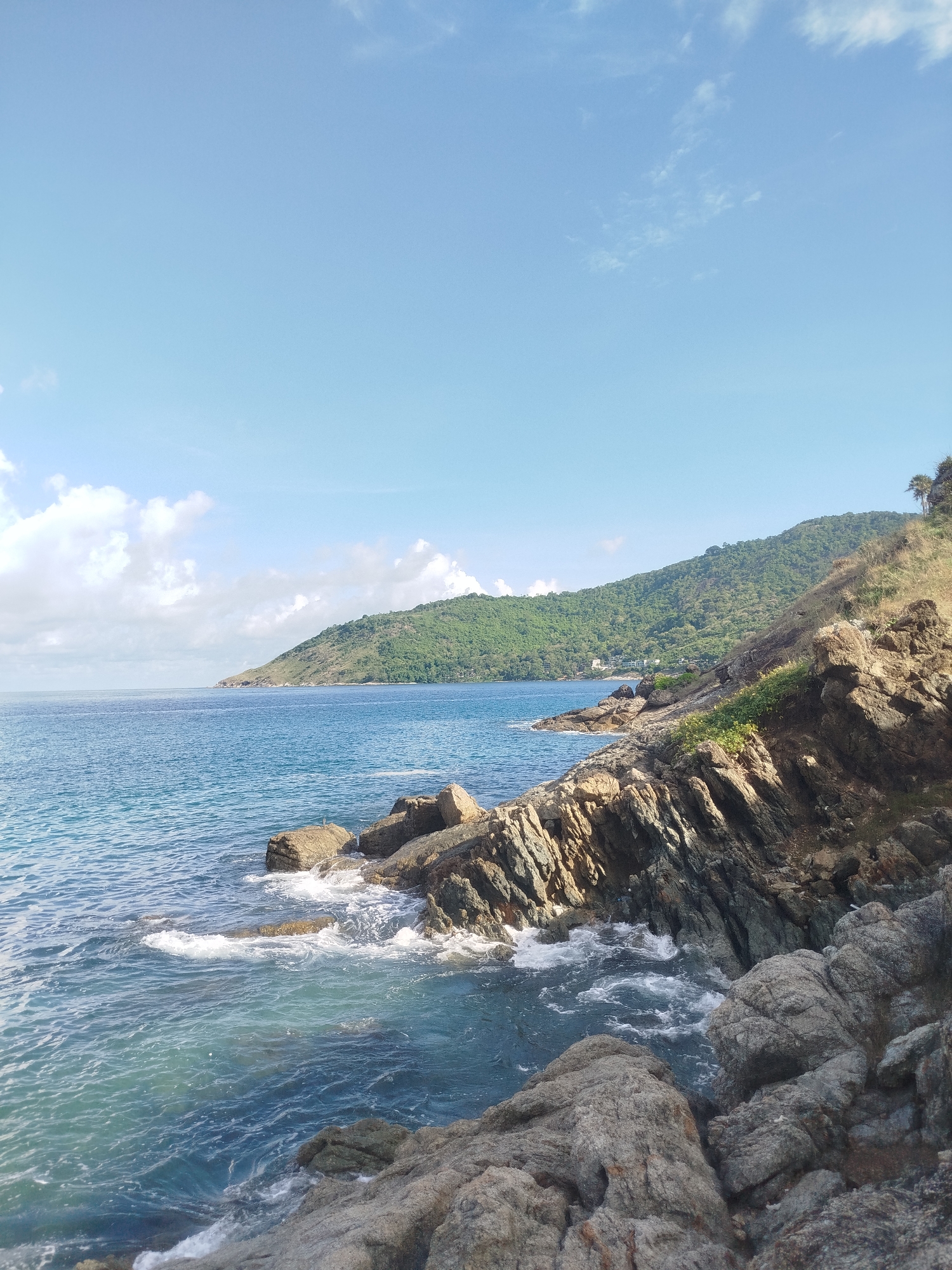 The image depicts a rocky coastline with the ocean and a lush green hill in the background. The sky is a clear blue with scattered white clouds. The foreground is dominated by rugged, textured rocks of various sizes, some of which are partially submerged in the turquoise water. Waves are gently crashing against the rocks, creating white foam. In the middle ground, the ocean stretches out, appearing calm and expansive. The background features a sloping hill covered in dense green vegetation, with a few buildings visible in the distance along the coastline. The scene is bathed in natural daylight, suggesting it was taken outdoors during the day. This view is consistent with coastal areas in Rawai, Thailand, known for their scenic beaches and hilly landscapes meeting the sea. There are no people or animals visible in the image.