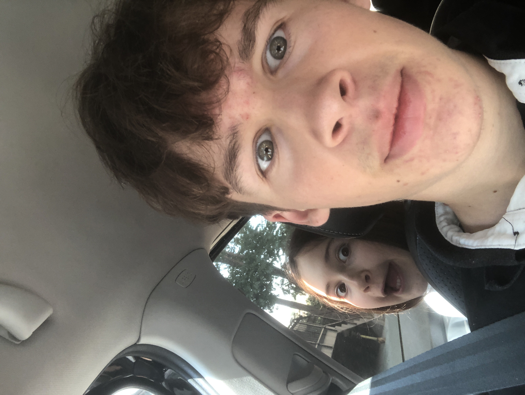 Two people, a male and a female, are in the interior of a vehicle, likely a car. The male is in the foreground, his face taking up most of the right side of the frame, looking directly at the camera. He has brown, messy hair and green eyes. The female is in the background, to the left of the male, with her face also directed towards the camera. She has brown hair and appears to be making a surprised or excited expression with wide eyes and an open mouth. Both individuals are wearing casual clothing. The interior of the car is visible, including part of the dashboard, an airbag label, and a seatbelt. Outside the vehicle, through the window, trees and a staircase are visible, suggesting an outdoor environment, possibly in a residential area. The lighting indicates it is daytime, with sunlight filtering through the trees. The image does not provide any specific information about the location being in Chișinău, Moldova.