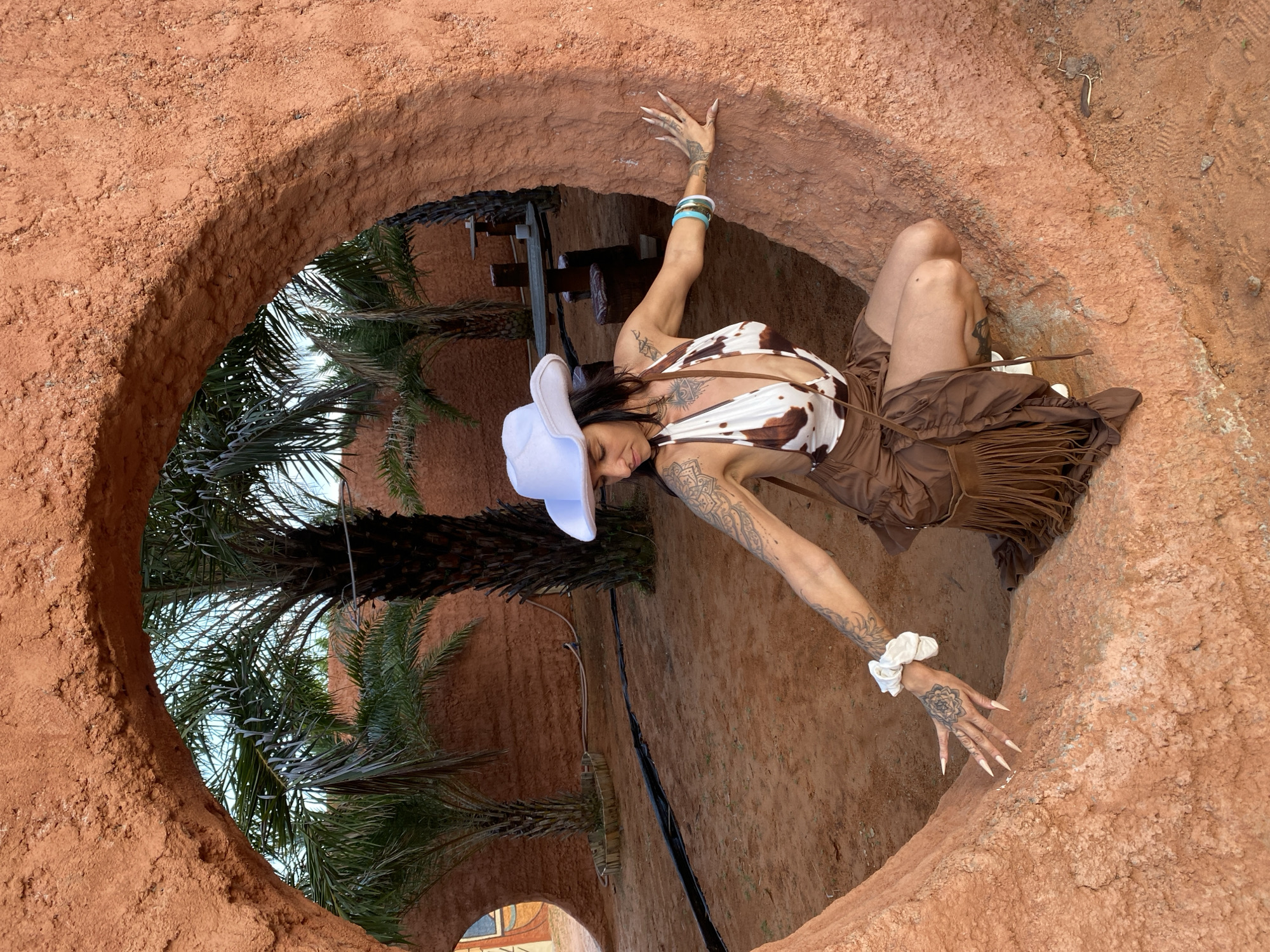 A woman wearing a white cowboy hat and a cow-print halter top is crouched inside a circular, rough-textured, terracotta-colored opening in a wall. She has long, pointed fingernails, tattoos on her arms and legs, and is wearing a brown skirt with fringe. She is leaning with her right hand on the wall and her left hand is extended towards the ground. In the background, palm trees and outdoor seating areas are visible under a blue sky, suggesting a desert or arid landscape, possibly within the city of Mui Ne, Vietnam, known for its sand dunes.