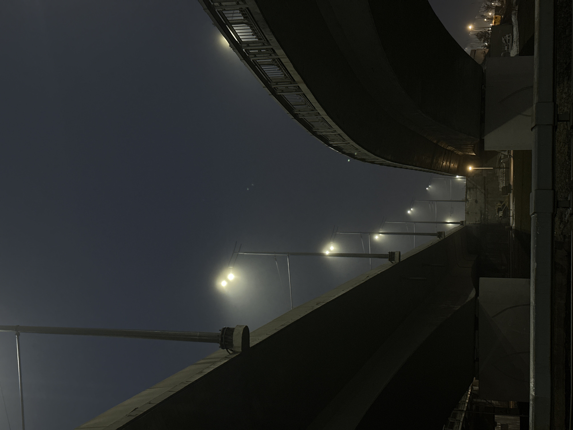 Captured from a low angle, the photograph showcases an urban scene at night in Kazan, Russia. Two curved bridge structures dominate the frame, with the space between them revealing a series of illuminated streetlights that recede into the distance. A faint glow emanates from these lights, hinting at foggy conditions. The bridge structures are primarily dark, with some reflective surfaces catching the light. The upper bridge features a distinct edge with a series of rectangular openings. The composition emphasizes the architectural elements and the interplay of light and shadow in the nighttime urban environment.