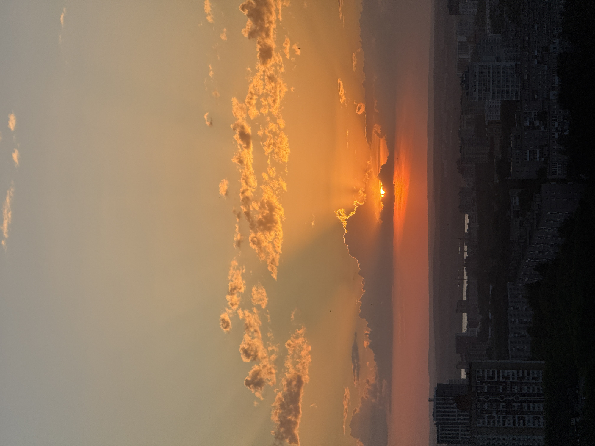 The image captures a cityscape at sunset, likely in Perm, Russia, given the presence of what appear to be typical urban high-rise buildings. The sun is partially obscured by clouds, with rays of light emanating from behind the cloud cover. To the left of the sun, a series of smaller clouds are present, illuminated by the sunlight. The sky gradually transitions from orange near the horizon to a lighter hue further up. The cityscape on the right is mostly silhouetted due to the low light, but the architectural details of the buildings are still somewhat discernible. There are no people immediately visible in the frame.