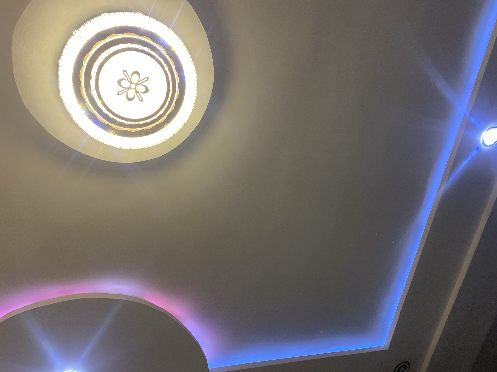 The image shows a modern ceiling with recessed lighting and a decorative chandelier. The ceiling is a neutral color, likely white or off-white, and features a stepped design with illuminated edges. A main chandelier with multiple concentric rings and a floral-like pattern in the center is prominently featured on the left side of the image. This chandelier emits a warm, bright light.
Along the right side of the ceiling, a strip of blue LED lighting creates a vibrant glow, outlining a recessed area. Towards the bottom left, another curved recess in the ceiling is illuminated with a pinkish-purple light, contrasting with the blue light on the right. The overall impression is one of contemporary interior design, likely in a home or a commercial space. The lighting suggests it could be evening or night, or simply that the artificial lighting is the primary illumination source. No people are visible in the image. The location context of Giginyu, Nigeria, does not provide specific visual cues within the image itself. There is no visible text.