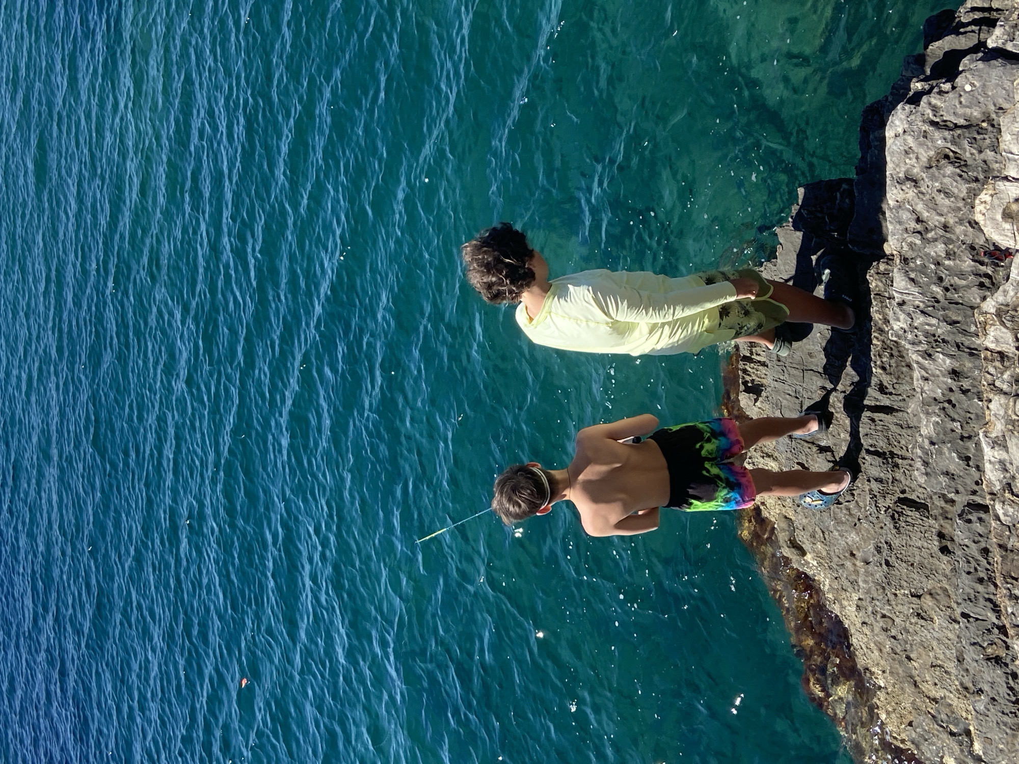 Two young males are standing on a rocky outcropping, looking out at the clear, blue-green water of the Adriatic Sea. The male on the left, facing away from the camera, is shirtless and wearing multi-colored swim trunks. He is holding a fishing rod and appears to be fishing. The male on the right, also facing away from the camera, is wearing a long-sleeved yellow shirt and light-colored shorts. He is looking down at the water. The scene is lit by bright sunlight, casting shadows on the rocks. The location is Budva, Montenegro, a coastal city known for its beaches and historic old town.