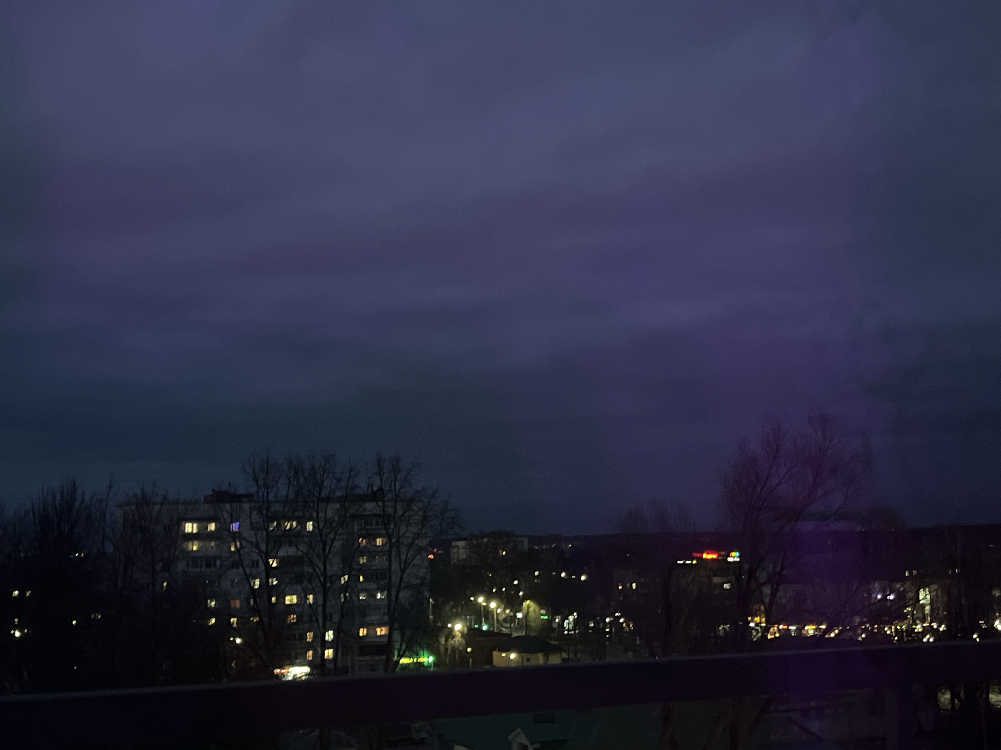 The image presents a nocturnal cityscape in Chaikovsky, Russia, viewed from an elevated position, likely a balcony or window. The dominant feature is the sky, which fills the upper two-thirds of the frame. It is a dark, overcast expanse, featuring visible cloud formations. The sky's color palette shifts from a deep, dusky blue on the left to a subtle, yet distinct, purplish tint on the right, possibly influenced by distant light sources or atmospheric conditions.
In the mid-ground, a multi-story apartment building stands prominently on the left. Many of its windows are aglow with warm, yellow light, signaling human presence and activity within. Bare, leafless deciduous trees, suggesting a winter or early spring season, are scattered throughout the mid-ground, partially obscuring the buildings.
Further into the distance, the urban landscape is dotted with numerous artificial lights. Bright streetlights cast distinct circular glows, illuminating parts of the streets and surrounding areas. Other distant buildings are visible, their forms indistinct against the darkness, but marked by scattered illuminated windows and commercial lights. A particularly bright, red-orange light, likely a commercial sign, appears in the upper right distance; however, no specific text is discernible.
The immediate foreground features a dark, horizontal railing or ledge, framing the urban vista. The overall atmosphere conveyed is one of quietude and stillness, typical of a city at night. No people or overt activities are visible; the scene captures a moment of static urban life after dark.