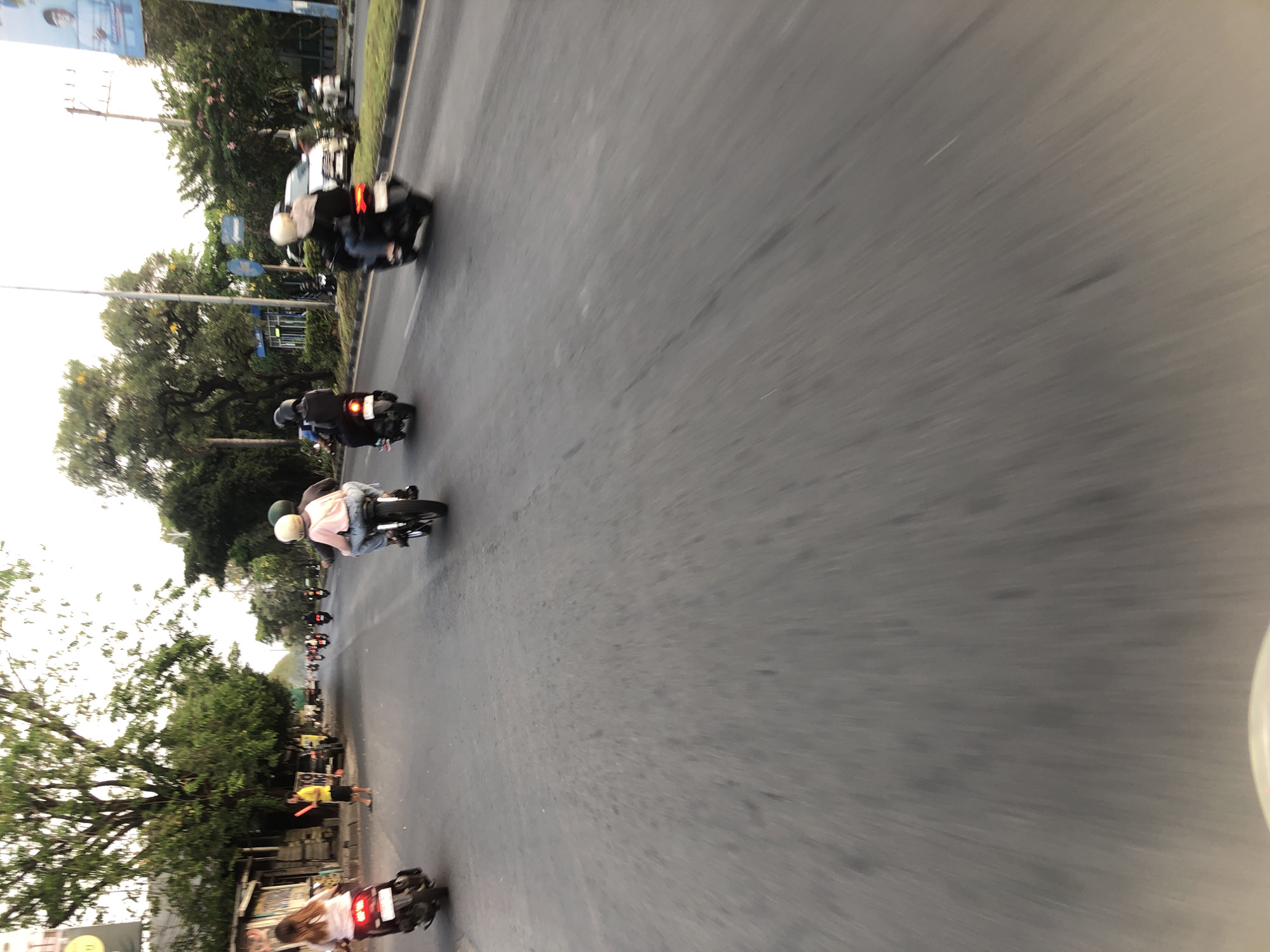In the city of Gelam, Indonesia, a group of people are riding motorbikes on a paved road. The scene appears to be during daylight, with a slightly overcast sky. The road has a white lane marker. On the left side of the road, there are several small buildings and trees. On the right side, there are more trees and a large billboard. The people on the motorbikes are wearing helmets and casual clothing. One motorbike has two people on it, while others have a single rider. The perspective of the image suggests it was taken from another moving motorbike. The overall impression is of everyday life in a busy urban or semi-urban environment.
