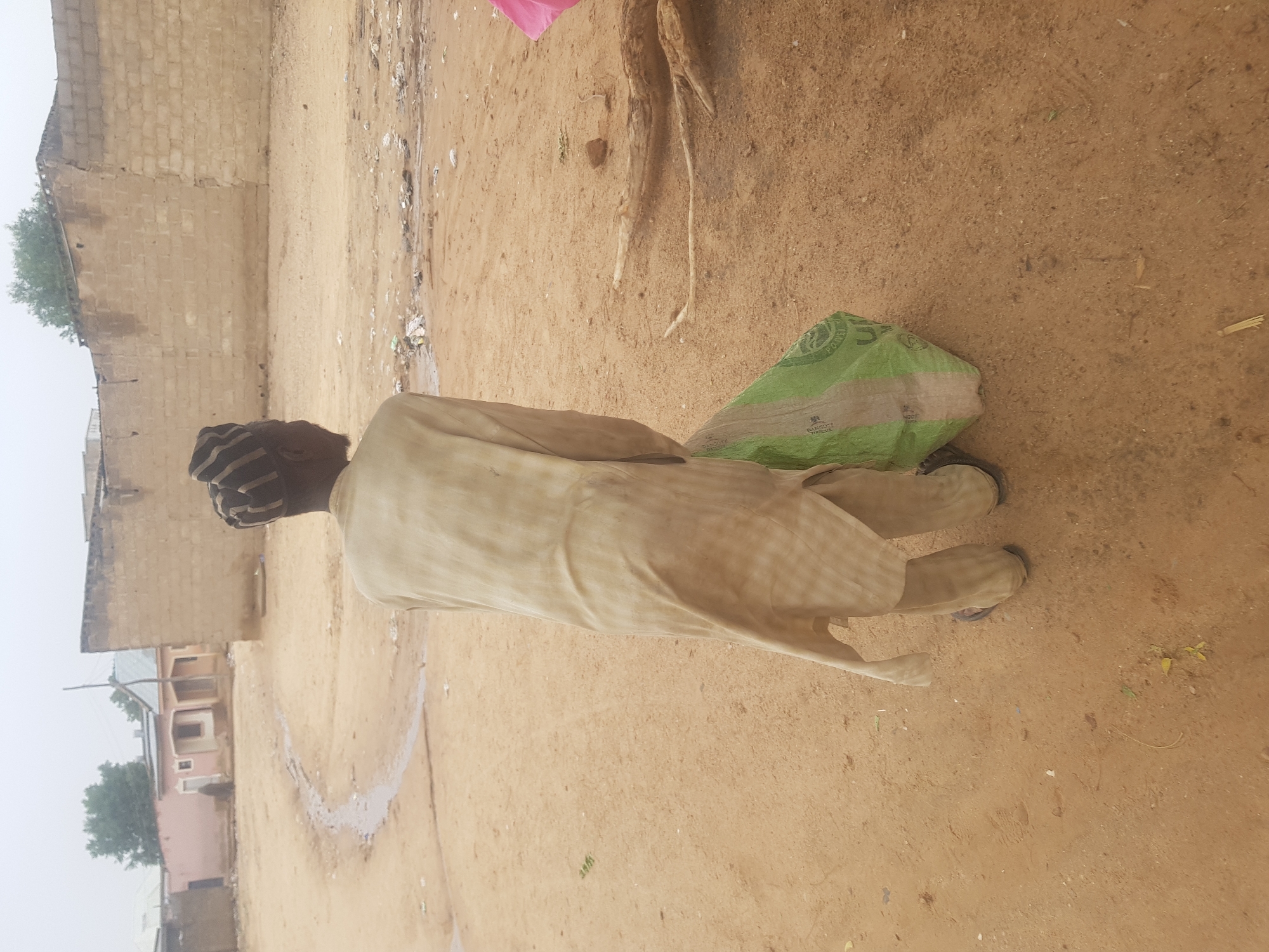An elderly person is standing in a sandy area in Dutsinma B, Nigeria, near a dirt road and some buildings. The person is wearing a long, light-colored garment and a striped cap. A green bag is on the ground near the person's feet. The environment appears to be a residential area, with a block wall and other buildings visible in the background. There is also some debris scattered on the ground.