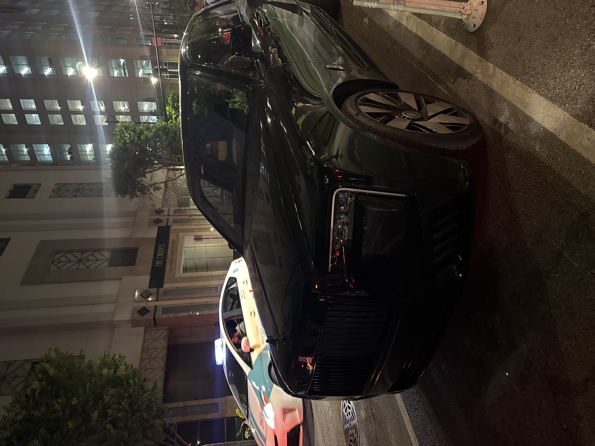 The image depicts a dark green Rolls Royce parked on a city street at night in Chicago, Illinois, USA. The front of the car is prominently displayed, with the distinctive grill and headlights visible. The car is positioned near the curb. A sign for "The Caravel" is visible on a building in the background. A taxi is also partially visible next to the Rolls Royce. The street is illuminated by streetlights, which reflect off the car's surface. The setting suggests a wealthy or upscale area of the city. The image suggests an urban environment.