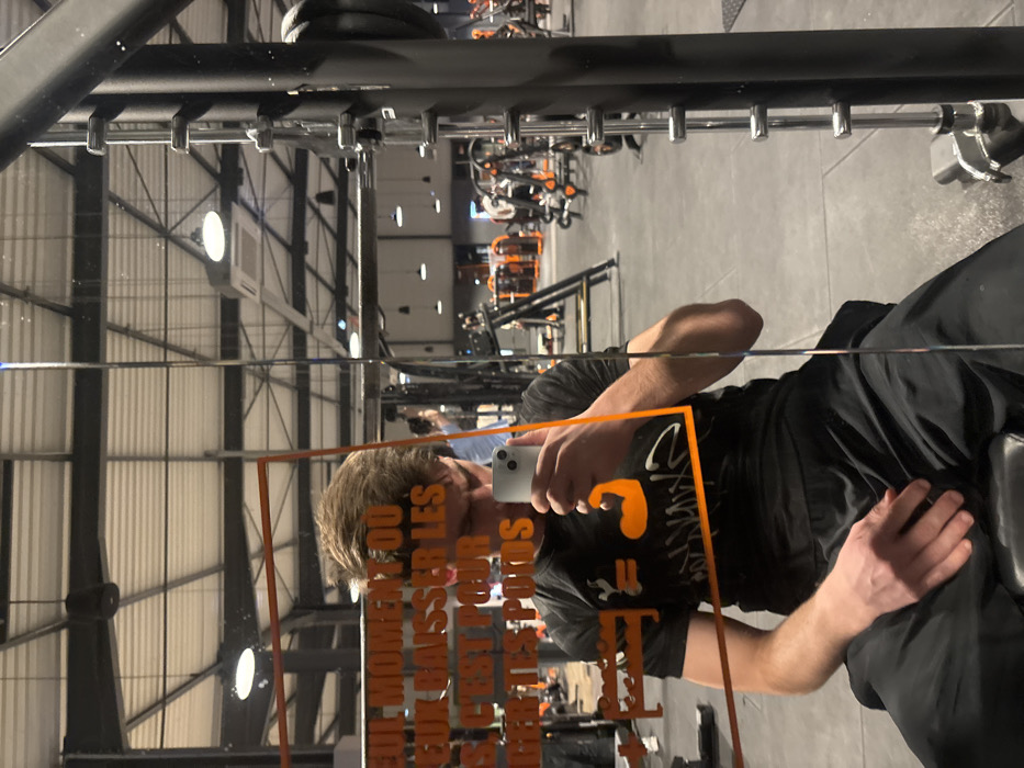 A selfie captured in a gym mirror reveals a person holding a phone, reflecting the camera. The gym's industrial ceiling with metal beams and overhead lighting is visible in the background. Weight training equipment, primarily in black and orange, is sparsely scattered throughout the gym. The person is wearing a black shirt and pants, and parts of a weight bench are reflected. The mirror has some orange text on it.