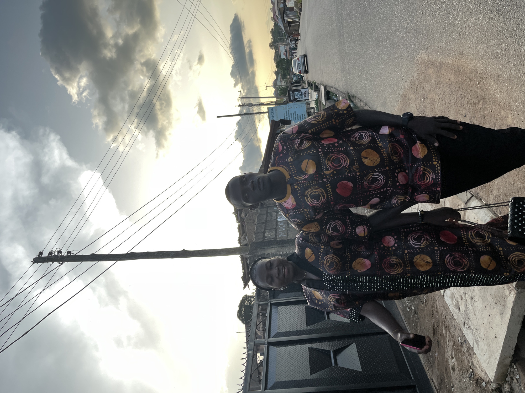 The image depicts two individuals, a man and a woman, walking on a sidewalk next to a paved road under an overcast sky at what appears to be late afternoon or early evening. The setting is consistent with an urban or semi-urban area in Nigeria, specifically requested as Jos.
The woman, positioned on the left, has a dark complexion and a slight smile. She wears a dark garment, possibly a long dress or tunic, featuring a pattern of abstract circles in shades of red, orange, and yellow, accented with vertical lines of white dots. She holds a pink mobile phone in her right hand and a small, dark, studded handbag in her left hand, which hangs by her side.
The man, on the right, also has a dark complexion and a serious expression, looking slightly to his right. He wears a short-sleeved top with the identical circular pattern as the woman's garment, suggesting matching attire. He wears dark trousers and a dark wristwatch on his left wrist.
They are walking side-by-side, moving towards the left side of the frame. Behind the woman, a black metal gate with a diamond plate pattern is visible, followed by a stone wall. A wooden utility pole with multiple overhead wires dominates the upper-center of the image, extending into the distance where more poles and wires are seen.
On the right side of the paved road, a street stretches into the distance, lined with low-rise buildings and what appear to be commercial establishments. A white sedan is visible on the road in the mid-ground. Prominent signs include one with blue text, partially visible as "B.P.C.C... COMPUTING SCHOOL," and another further down displaying "THE GREAT POS" with a graphic of a hand. The ground on the sidewalk is uneven concrete and dirt, with a visible drainage channel running parallel to the road. The sky is largely covered with dark, heavy clouds, but bright light breaks through from the upper left, indicating a low sun.