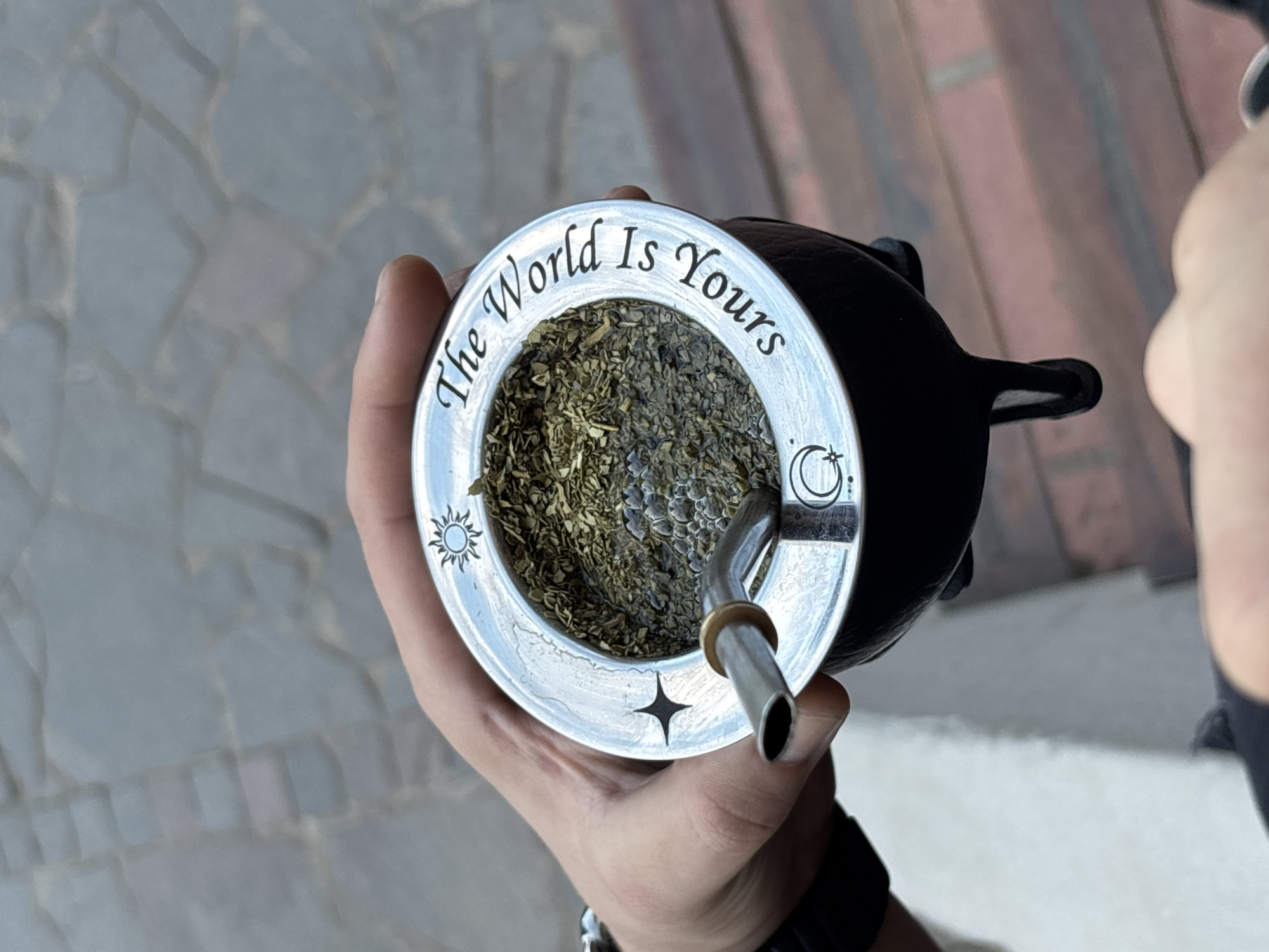 A person holds a mate gourd filled with yerba mate and a bombilla (straw) in their hand. The gourd has a silver rim with the phrase "The World Is Yours" engraved around the edge, accompanied by small sun, star, and moon symbols. The background suggests an outdoor setting, with stone pavement visible. Based on the context of a mate gourd, the location may potentially be Villa General Belgrano, Argentina.