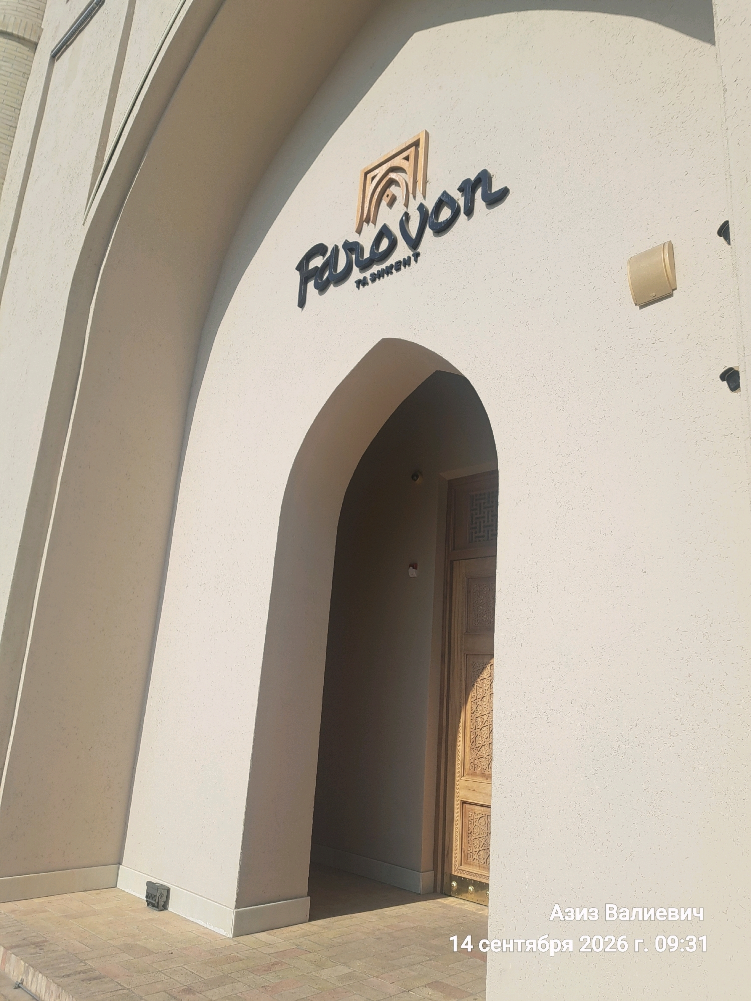 The image shows the exterior of a building with a large arched entrance in Tashkent, Uzbekistan. The building's facade is a textured beige color. Above the arch, a sign reads "Farovon" in dark lettering, with "TASHKENT" below it. The logo above "Farovon" is a stylized arch. To the right of the sign, a cream-colored rectangular speaker is mounted on the wall. The archway leads to a wooden door with intricate carvings. In the lower right corner of the image, text in Russian reads "Азиз Валиевич 14 сентября 2026 г. 09:31", indicating the name Aziz Valievich and the date September 14, 2026, at 09:31. The ground in front of the entrance is paved with brick. The lighting suggests it is daytime, with shadows cast by the building's architecture.