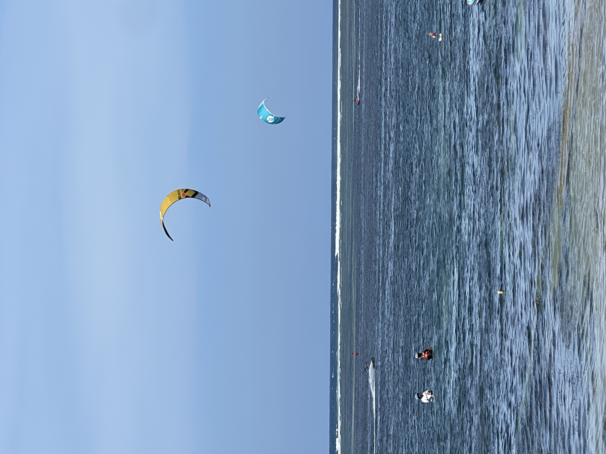 This media file shows a kitesurfing scene on a sunny day. The image is oriented vertically, with the sky on the left and the ocean on the right.
Two kites are visible in the sky. The larger kite, in the foreground, is yellow with black and red accents and is shaped like a crescent moon. Further back, a smaller, blue kite with white designs is also in the air.
The ocean is a deep blue with whitecaps, indicating a moderately choppy surface. Several people are engaged in kitesurfing. One person in a red vest is clearly visible on the water, while other individuals can be seen as smaller figures on surfboards further in the distance.
The sun is high in the sky, suggesting it is midday. The overall mood is one of activity and recreation, with people enjoying watersports under clear skies. No specific location cues other than the general beach and ocean setting are apparent within the image itself.