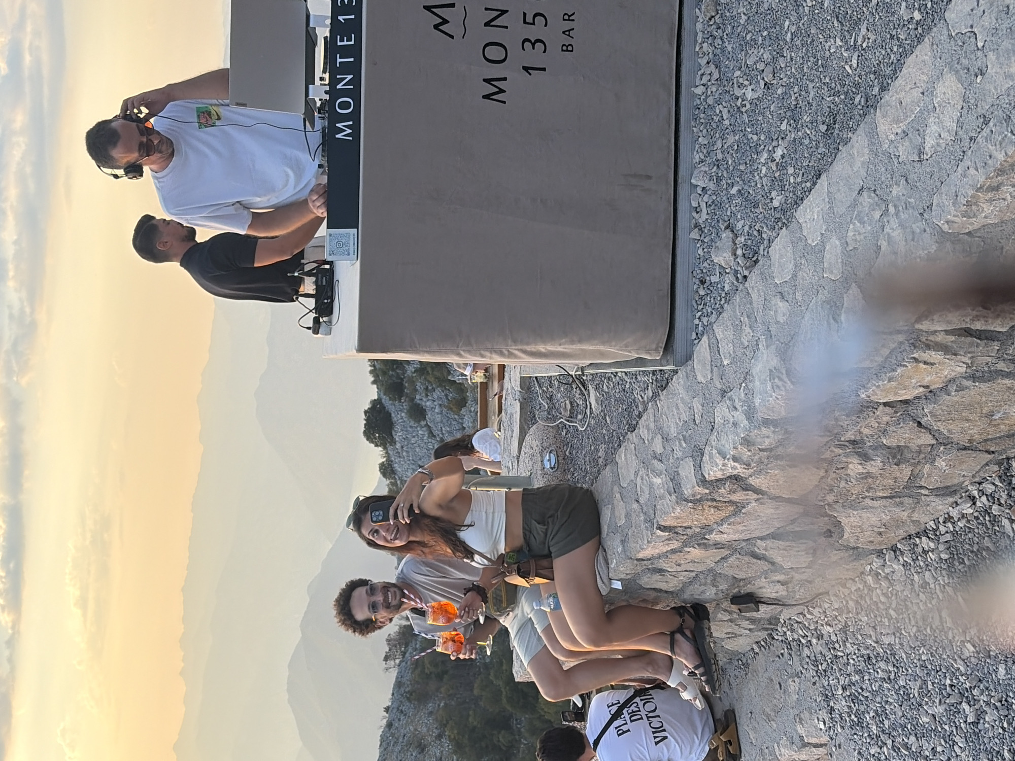 A group of people are gathered at a bar on a mountainside in Kuk, Montenegro, during sunset. The sky is a warm orange and yellow, with layers of hazy mountains in the background. In the foreground, a couple is seated on a stone wall, both holding drinks and smiling. The woman is taking a selfie with her phone. To the right, a DJ is playing music, with another person standing behind him. A beige bar with "MONTE 1350 BAR" printed on it is visible. In the background, other patrons are seated at tables. The ground is covered in gravel and small stones.