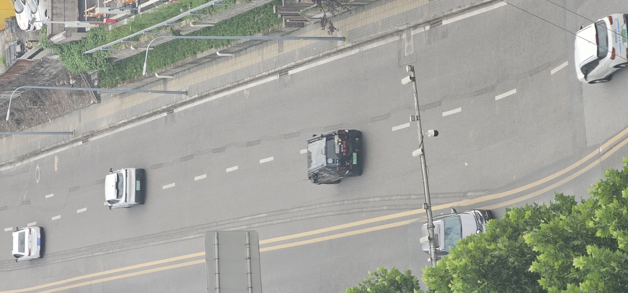 The image shows an aerial view of a street in Jiulongpo District, China. In the foreground, a silver SUV is parked on the left side of the road, partially obscured by lush green trees. A traffic camera gantry extends across the road, equipped with multiple cameras. Further down the road, a black SUV is driving in the right lane. The road has double yellow lines marking the lanes and white dashed lines indicating lane divisions. To the right of the road, a sidewalk runs alongside a retaining wall covered in vegetation, with lampposts and a fence. Further up the road, two white cars are visible, one in the left lane and another in the right lane. A white car is also seen at the bottom right of the frame. The scene depicts a typical urban street environment with moving and parked vehicles.