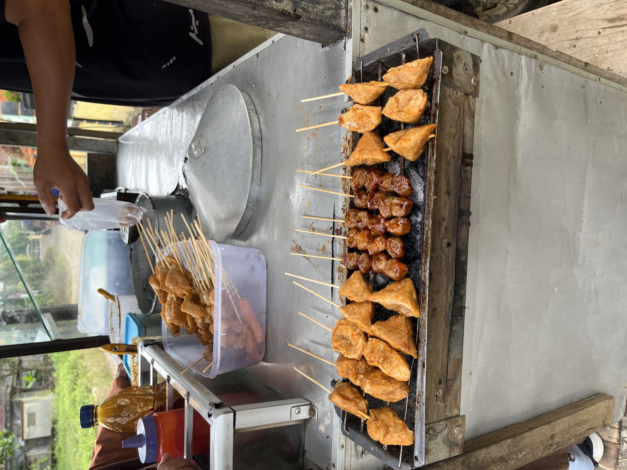 The scene depicts a street food vendor's cart in Matangpanyang, Indonesia, where various skewered items are being grilled and prepared outdoors during the daytime.
In the foreground, a metal charcoal grill contains multiple skewers of food. On the left side of the grill, approximately twelve skewers hold light orange-brown, irregularly shaped fried items, some triangular and some rounded, suggestive of fried tofu or fish cakes. On the right side, about ten skewers bear smaller, darker brown, glazed, spherical items, likely meatballs or similar protein skewers. Black charcoal embers are visible beneath the grill rack.
Above the grill, a metal cart forms the primary workspace. On the left side of the cart's metal countertop, a clear plastic bottle with a blue cap contains a thick, yellowish-brown liquid. Adjacent to it is a red squeeze bottle with a purple nozzle, presumably holding chili sauce. A clear plastic rectangular container holds numerous ungrilled or partially prepared skewered food items, similar to those on the grill, along with a few longer, reddish items (possibly sausages). Several other metal and plastic containers, some with lids (one light blue, one white), are arranged on the countertop.
On the right side of the countertop, a large, conical metal lid rests, possibly covering a pot or steamer. A person, visible from the mid-torso to the upper arm, wearing a black t-shirt with white patterns, is holding a small, clear plastic bag containing food items over a metal bucket or container. Another person's arm, dressed in a brown sleeve, is partially visible on the far left, near the sauce bottles. Their lower leg and a black shoe with a white sole, worn with a blue and white striped sock, are also visible at the bottom left.
The background, out of focus, shows blurred greenery consistent with trees and grass, along with indistinct structures or buildings, indicating an outdoor or roadside setting. The ground beneath the cart is a light-colored surface, possibly concrete, with some wooden planks or supports visible at the edges.
