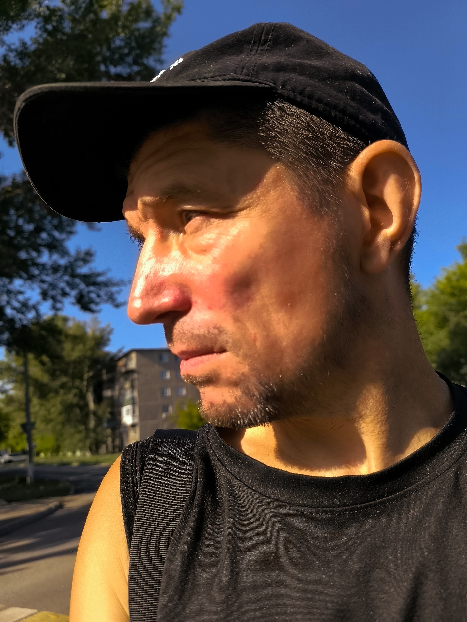 A man wearing a black baseball cap and a black t-shirt is pictured in a close-up profile shot. The man's face is illuminated by harsh, golden sunlight, creating strong shadows and highlights. He has dark hair, stubble on his chin, and a reddish hue on his nose and cheeks. The background consists of a bright blue sky, green trees, and a multi-story building. A road with a yellow curb is visible in the lower left of the frame, suggesting an urban or suburban environment. The scene appears to be outdoors, possibly in a park or along a street in Qarağandı city administration, Kazakhstan, given the presence of Soviet-era architecture.