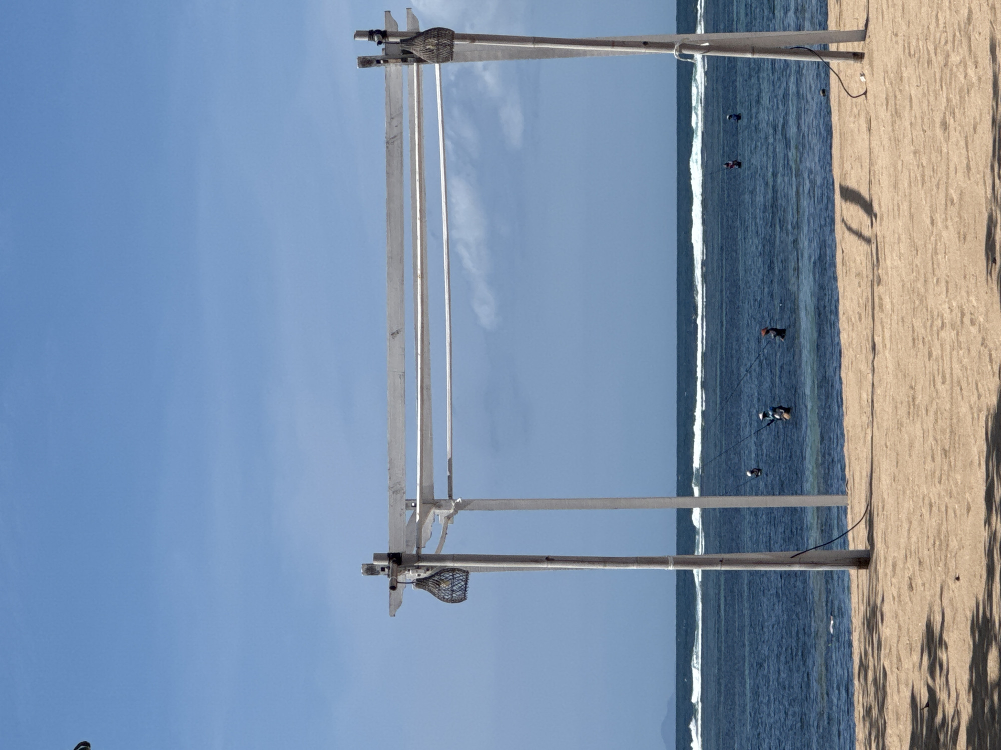 The image depicts a beach scene in Kuta Selatan, Indonesia. A metal structure stands on the sandy beach, adjacent to the ocean. The structure comprises vertical posts and horizontal bars, topped with hanging, netted baskets. In the background, the ocean stretches to the horizon, with visible waves. Several surfers are visible in the water. The sky above is clear and blue. The beach is sandy with shadow patterns cast by unseen elements.