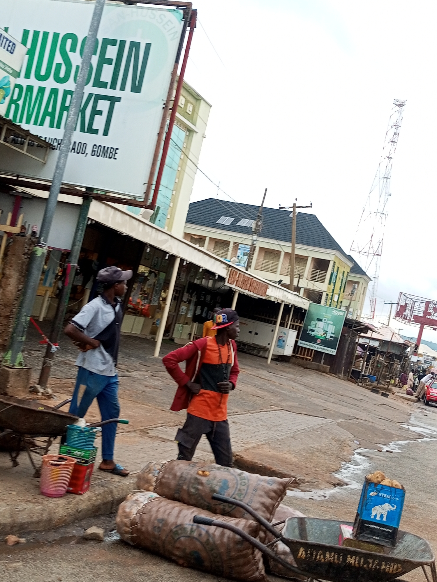 The image depicts a street scene in Arawa, Nigeria. In the foreground, two men are visible. One man, wearing a grey shirt, black tie, and blue jeans, stands looking to the right with his hand on his hip. Another man, wearing a red jacket over an orange shirt and dark pants, stands further to the right, also looking to the right.
In front of the men, there is a wheelbarrow filled with sacks. To the left of the wheelbarrow are stacks of colorful containers, including a pink bucket, green baskets, and red crates.
In the background, there is a large billboard with green lettering that reads "HUSSEIN MARKET". Adjacent to the billboard is a multi-story building with light-colored walls and dark-tiled roofs. Further in the background, a tall radio tower is visible against an overcast sky. Other stalls and buildings line the street, and a red car is partially visible in the distance. The ground is paved with asphalt and has some patches of water or oil.