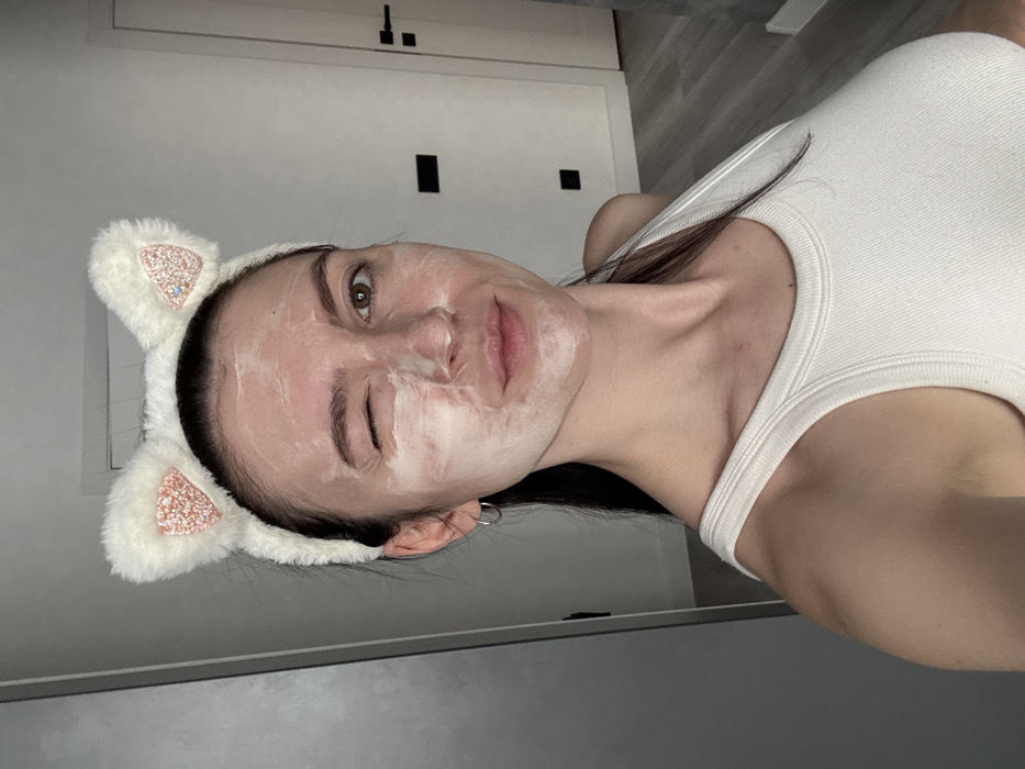 The image captures a selfie-style portrait of a young woman, rotated 90 degrees clockwise. When viewed upright, she is looking directly at the camera with a playful expression. She is winking her right eye while her left eye is open and appears to have a slight pout or soft smile on her lips.
Her face is covered in a white, creamy substance, likely a face mask or skincare product, which is spread unevenly across her cheeks, forehead, and nose, indicating she is in the midst of a beauty or skincare routine.
She is wearing a white, fluffy headband adorned with cat ears. The inner sections of the cat ears are covered in sparkling glitter, which appears to be a light pink or rose gold color. Her dark hair is pulled back by this headband.
She is dressed in a simple white ribbed tank top. A small, thin silver hoop earring is visible in her left earlobe.
The background is an indoor setting, appearing to be a light-colored wall or door, possibly off-white or light grey. Two dark rectangular objects, resembling light switches or electrical outlets, are visible on the wall behind her head. A slightly darker, greyish area is visible towards the bottom of the frame (when upright), which could be a floor or a piece of furniture. The lighting is soft and even, suggesting an indoor environment without harsh shadows. No text is visible in the image.